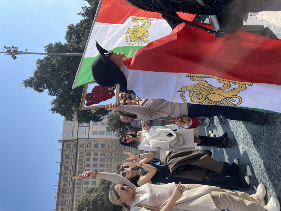 A daytime outdoor gathering is depicted on Plaça de Catalunya in Barcelona, Spain. Approximately 10-15 individuals, primarily women, are visible. In the foreground, a woman wearing a black wide-brimmed hat is draped in a historical Iranian flag (green, white, red, with a golden Lion and Sun emblem), seen from her right profile with her head turned to the right. To her left, another woman, wearing a beige top and sunglasses, holds a framed mirror containing a red, floral, map-shaped representation of Iran. Further left, a woman in a light beige outfit and sunglasses raises her right arm, index finger extended upwards. Nearby, another woman in a black outfit claps her hands. Several other participants are visible in the mid-ground, some smiling, others clapping. The ground is dark, patterned paving. Strong sunlight casts clear shadows under a clear blue sky. In the background, large trees are visible, along with a multi-story building featuring a stone facade and numerous windows, characteristic of central Barcelona architecture, on the left, and a more modern building partially visible behind it. A street light pole is present in the upper right.