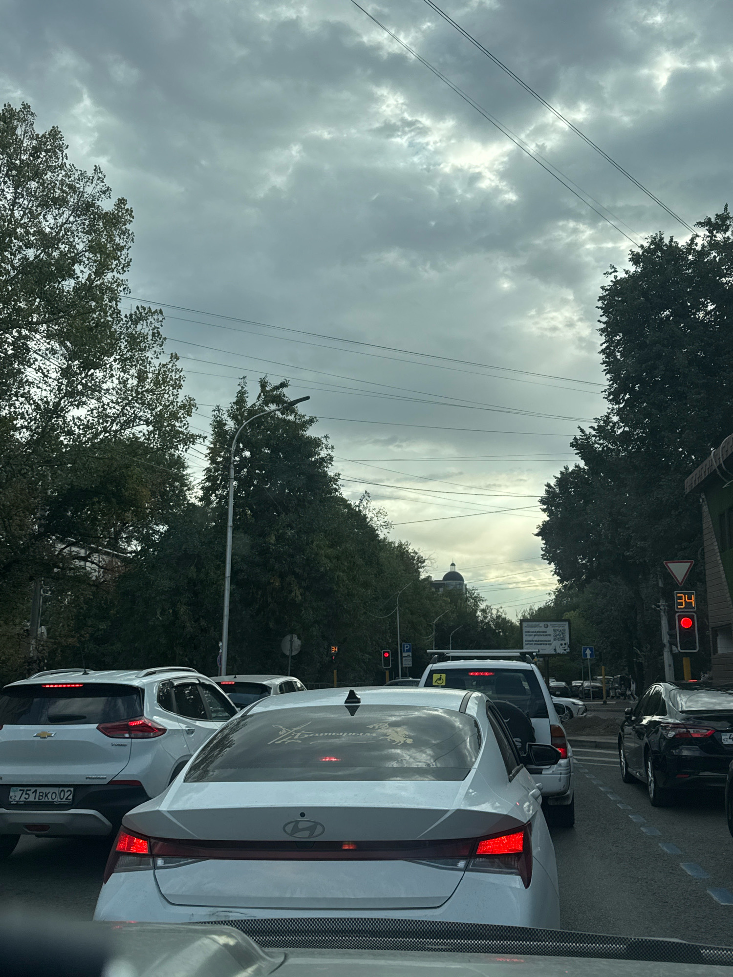 The image depicts a traffic jam on a city street in Almaty, Kazakhstan, under an overcast sky. Several cars are stopped in traffic, including white sedans and SUVs, and a black sedan. A traffic light shows red, and a digital display reads "34". Trees line the street, and power lines are visible overhead. In the distance, a building with a dome is visible. The license plate of one of the white SUVs reads "KZ 751BKO 02".