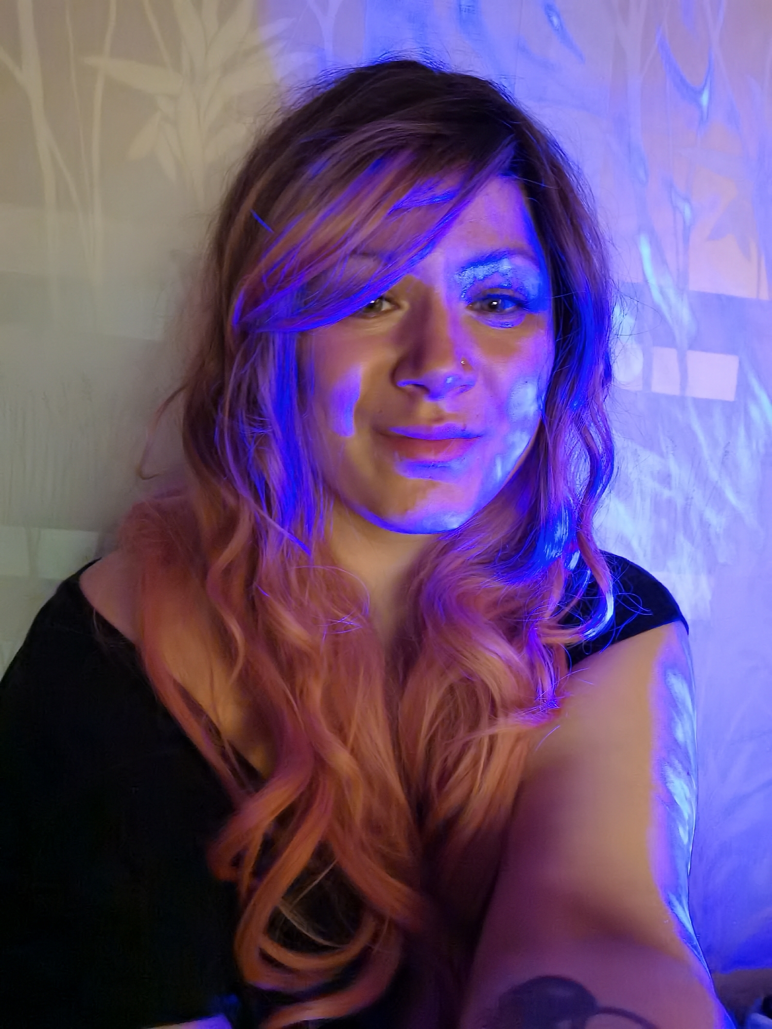 A woman with long, wavy, auburn and brown hair is centrally framed in a close-up shot. She is wearing a black top, and her right arm, bearing a tattoo, is visible in the foreground. Her face is illuminated with blue light, which also casts patterns on the wall behind her. The backdrop appears to be a textured or patterned wall, possibly wallpaper. No information suggests a location in Chaikovsky, Russia.