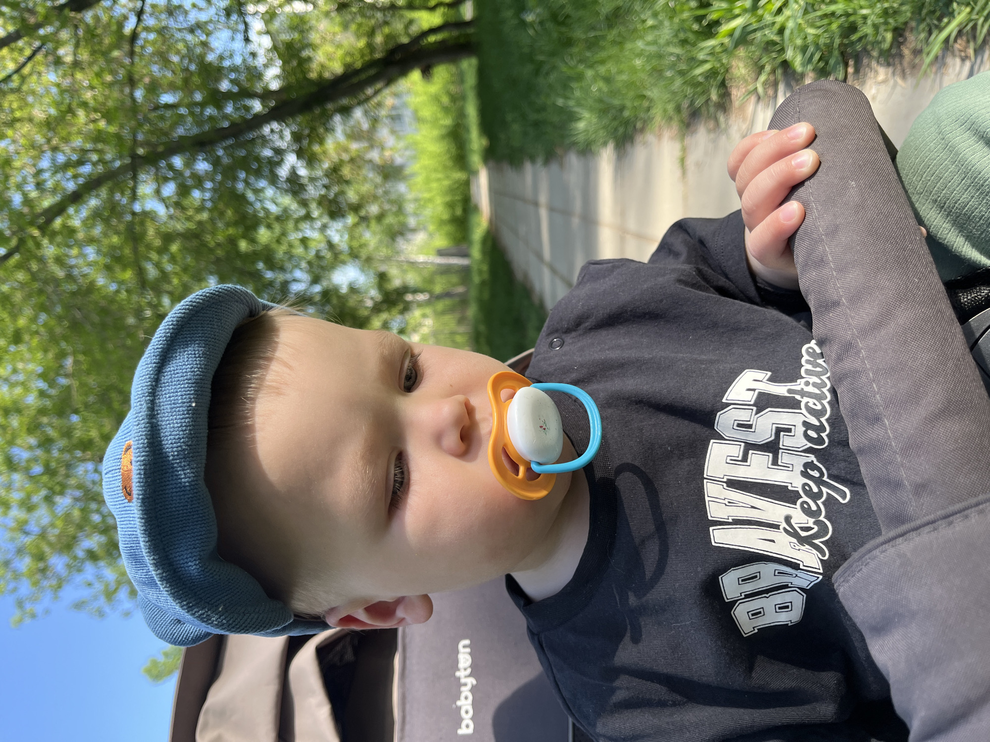 A young child is sitting in a stroller outdoors. The child wears a blue knit cap and a black t-shirt with white lettering. The child has a pacifier in their mouth and holds onto the gray padded bar of the stroller with their right hand. In the background, there is a sidewalk, greenery, and trees. The location is Chaikovsky, Russia.