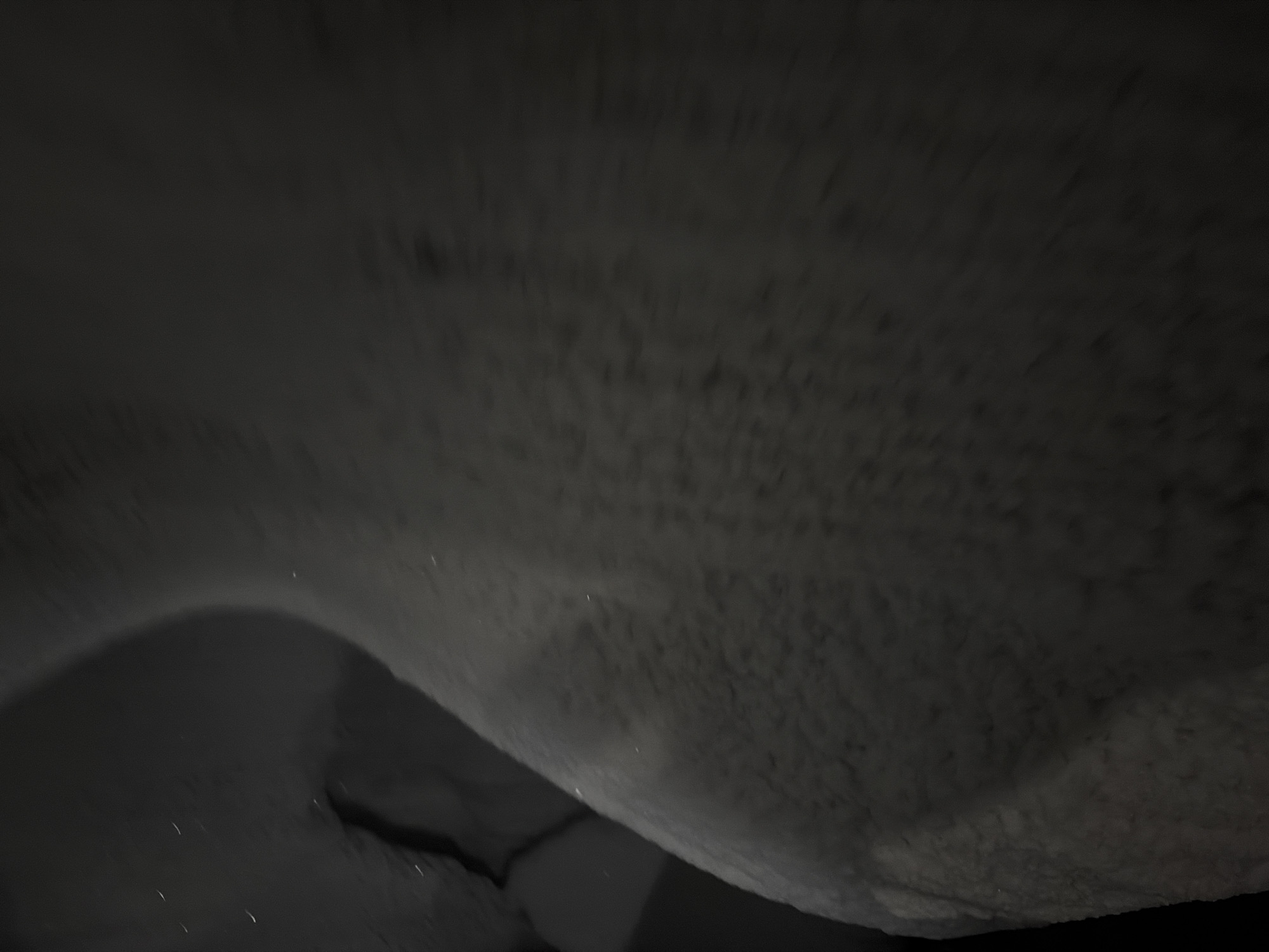 The image is a close-up, low-angle shot of a snow-covered surface, likely outdoors. The snow appears to be textured with small, granular formations, suggesting it may have been recently fallen or wind-swept. There are a few small, bright specks that could be ice crystals or other reflective particles within the snow. The lighting is dim, creating deep shadows and a muted color palette of grays and blacks. The perspective is such that the surface seems to curve upwards and away from the viewer. No specific location in Moscow, Russia, can be identified from this abstract and unidentifiable natural element.
