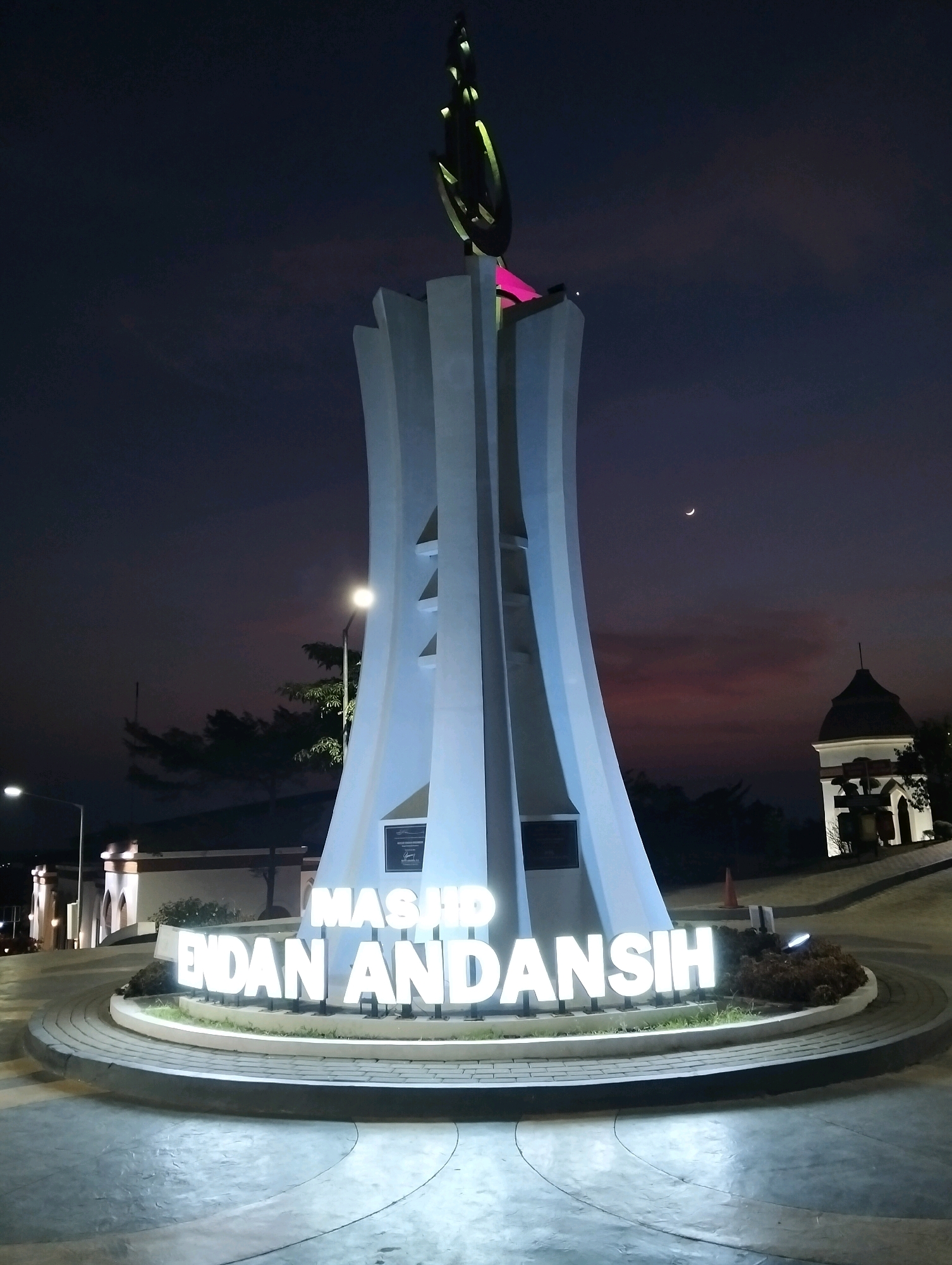 A monument with illuminated text reading "MASJID ENDAN ANDANSIH" stands in a public space at night. The monument is a tall, white structure with a curved, tapering design, topped with a decorative Islamic symbol illuminated in green and pink. The text is rendered in large, white, three-dimensional block letters that are also illuminated. The scene is set against a dark, twilight sky showing a crescent moon and some wispy clouds tinged with pink. To the right of the monument, a small, white, domed building with arched windows is visible, also illuminated. The foreground shows a paved area with a circular, stepped design. Trees and streetlights are also present in the background. This monument is located in the city of Purwakarta, Indonesia. There are no people visible in the image.