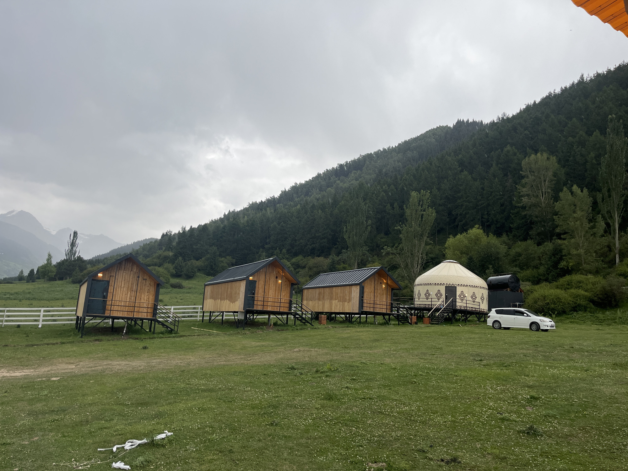 In the city of Karakol, Kyrgyzstan, an overcast day presents a scenic landscape featuring a glamping site. In the foreground, a grassy field stretches towards a row of three identical wooden cabins, each elevated on stilts and accessible by stairs. To the right of the cabins, a traditional yurt stands, adorned with ornate patterns. A white car is parked near the yurt. Behind the structures, a dense, dark green forest covers a sloped mountain that rises towards a cloudy sky. A white fence runs along the left side of the field.