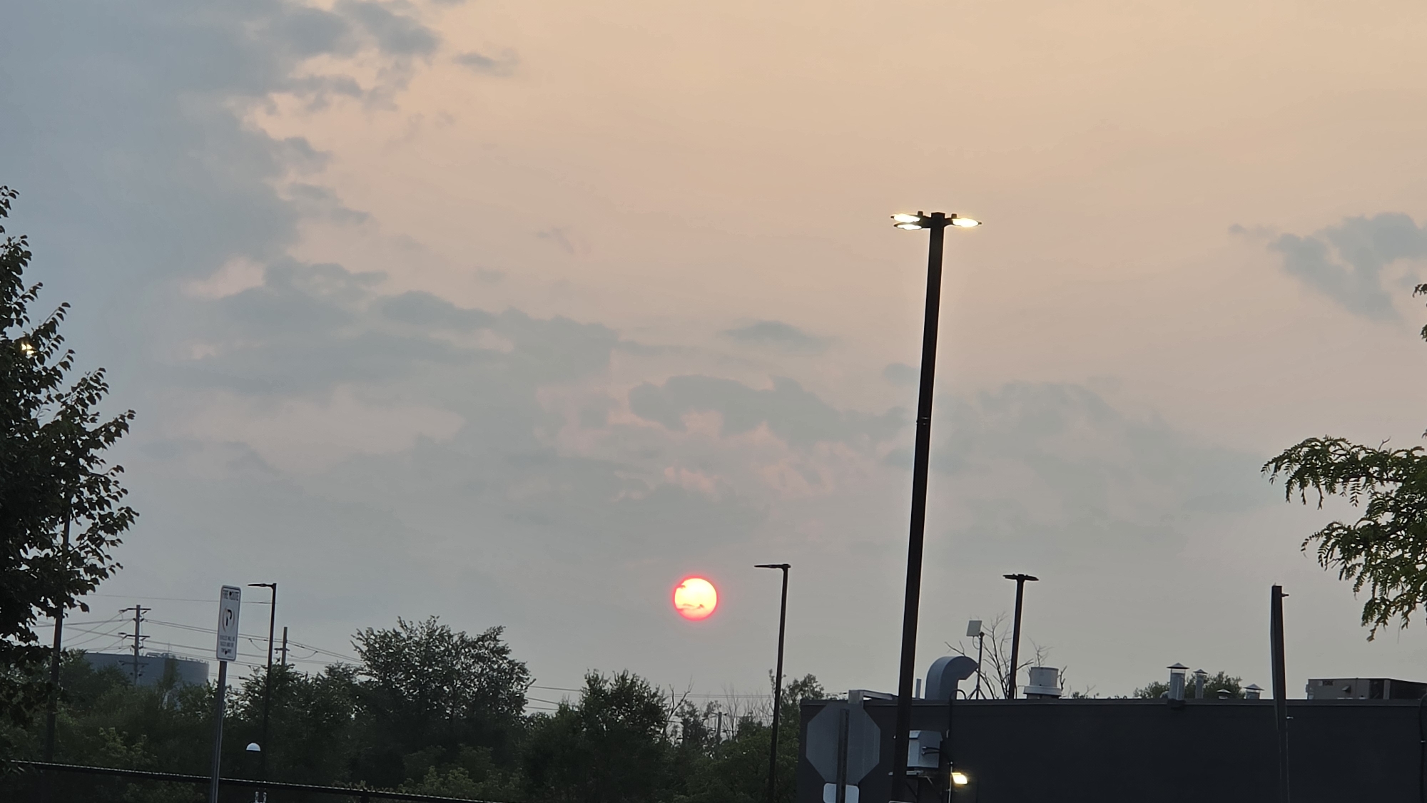 This image captures a sunset over an urban landscape in Markham, Canada. The sun, a bright red orb, is partially obscured by clouds, casting a soft glow across the sky. Several tall lampposts, some illuminated, punctuate the scene, suggesting an outdoor public space or commercial area. In the foreground, trees line a fence, with parts of buildings, including rooftops and ventilation systems, visible in the mid-ground. Utility poles and wires are also present, indicating the infrastructure of an urban environment. The overall atmosphere is one of twilight, with the fading light creating a somewhat muted color palette.