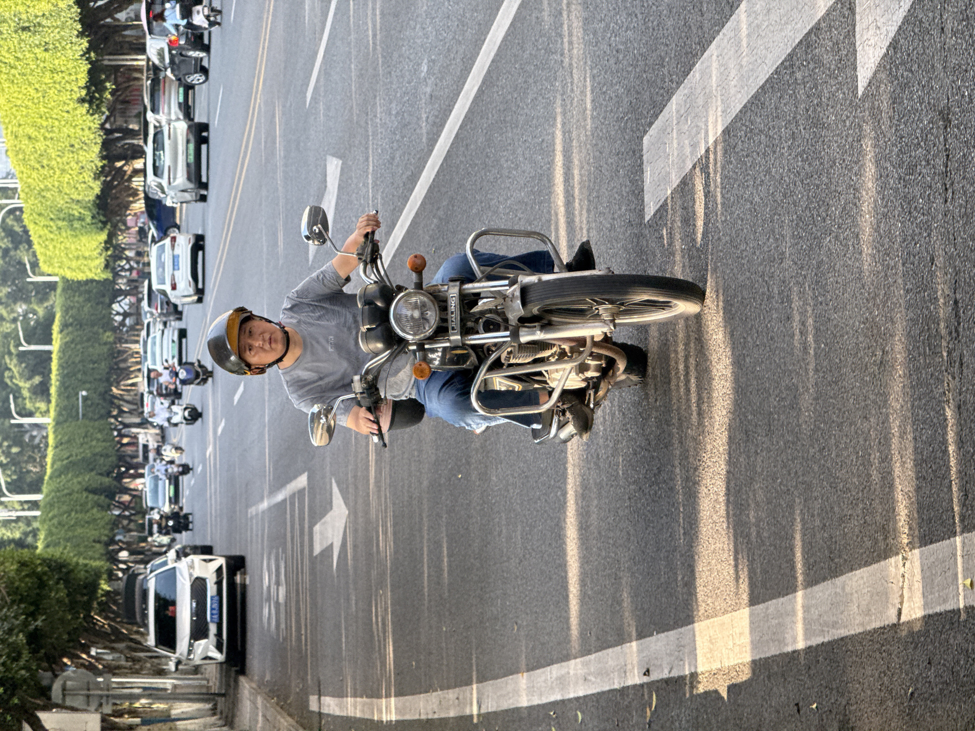 An eye-level shot captures a person on a motorcycle on a multi-lane asphalt road during the late afternoon or early evening, indicated by the long shadows and golden light. The image is taken from a slightly elevated perspective, looking down the road.
In the foreground, a person wearing a grey t-shirt, blue jeans, and a brown and gold helmet is seated on a classic-style motorcycle. They are looking towards the camera with a neutral expression. The motorcycle features chrome accents, a prominent round headlight, and visible rearview mirrors. The brand "JIALING" is clearly legible on the front frame of the motorcycle, below the headlight.
The road features clear white lane markings, including solid lines, dashed lines, and a large white arrow indicating direction of travel. In the background, a line of traffic is visible, consisting of various cars (including a white SUV with a partially visible license plate "A 8L2V6") and other motorcycles or scooters. The left side of the road is bordered by a continuous line of neatly trimmed green bushes or trees, creating a treelined streetscape. The overall scene depicts a busy urban or suburban street in Jiangjin, China, with vehicles moving or waiting in traffic under sunny weather conditions.
