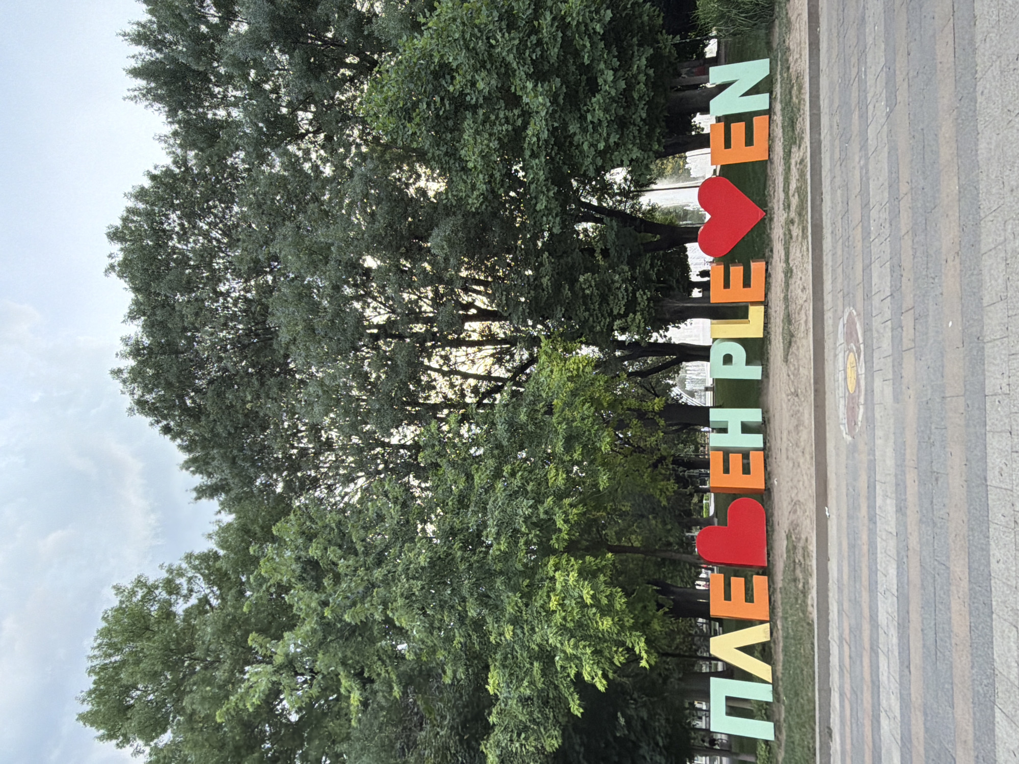A large, colorful sign spelling out "PLEVEN" with two heart shapes replacing letters is the central focus of the image. The sign is situated in a park-like setting with lush green trees in the background. In the distance, a fountain can be seen. The foreground shows a paved walkway with a patterned design. The sky is overcast with some clouds visible. The overall scene depicts a public space in the city of Pleven, Bulgaria, likely a landmark or tourist attraction.