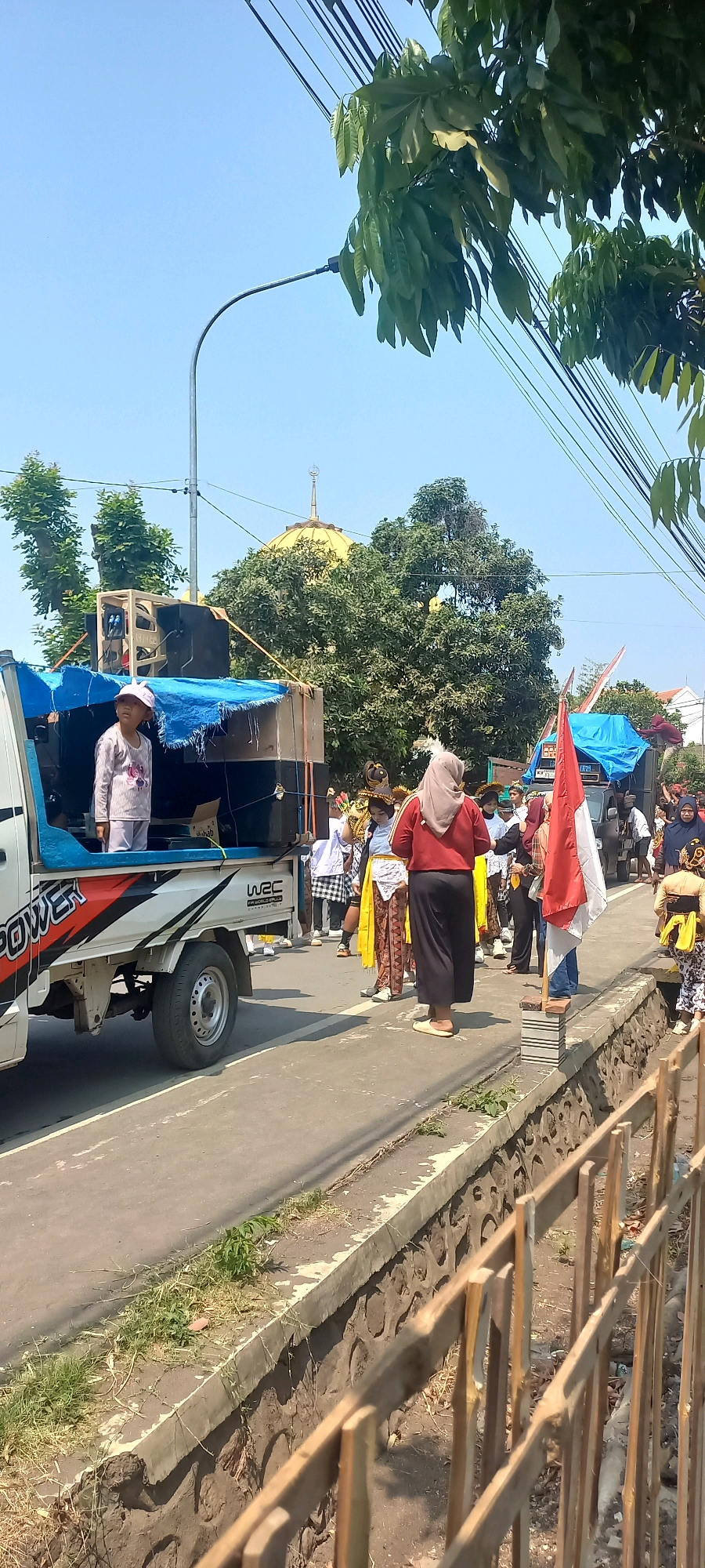 In Tongaswetan, Indonesia, a procession is underway on a paved road lined with a stone and concrete retaining wall and a wooden fence. A white pickup truck, decorated with "POWER" and "WRC" graphics, carries large speakers and is covered with a blue tarp. A young girl in a pink hat stands on the truck bed. Several individuals in traditional attire, including women in headscarves and men in patterned skirts and elaborate headdresses, are visible. One person holds a red and white Indonesian flag. In the background, lush green trees surround a golden-domed building, possibly a mosque, under a clear blue sky. The scene suggests a cultural event or celebration taking place outdoors during the day.