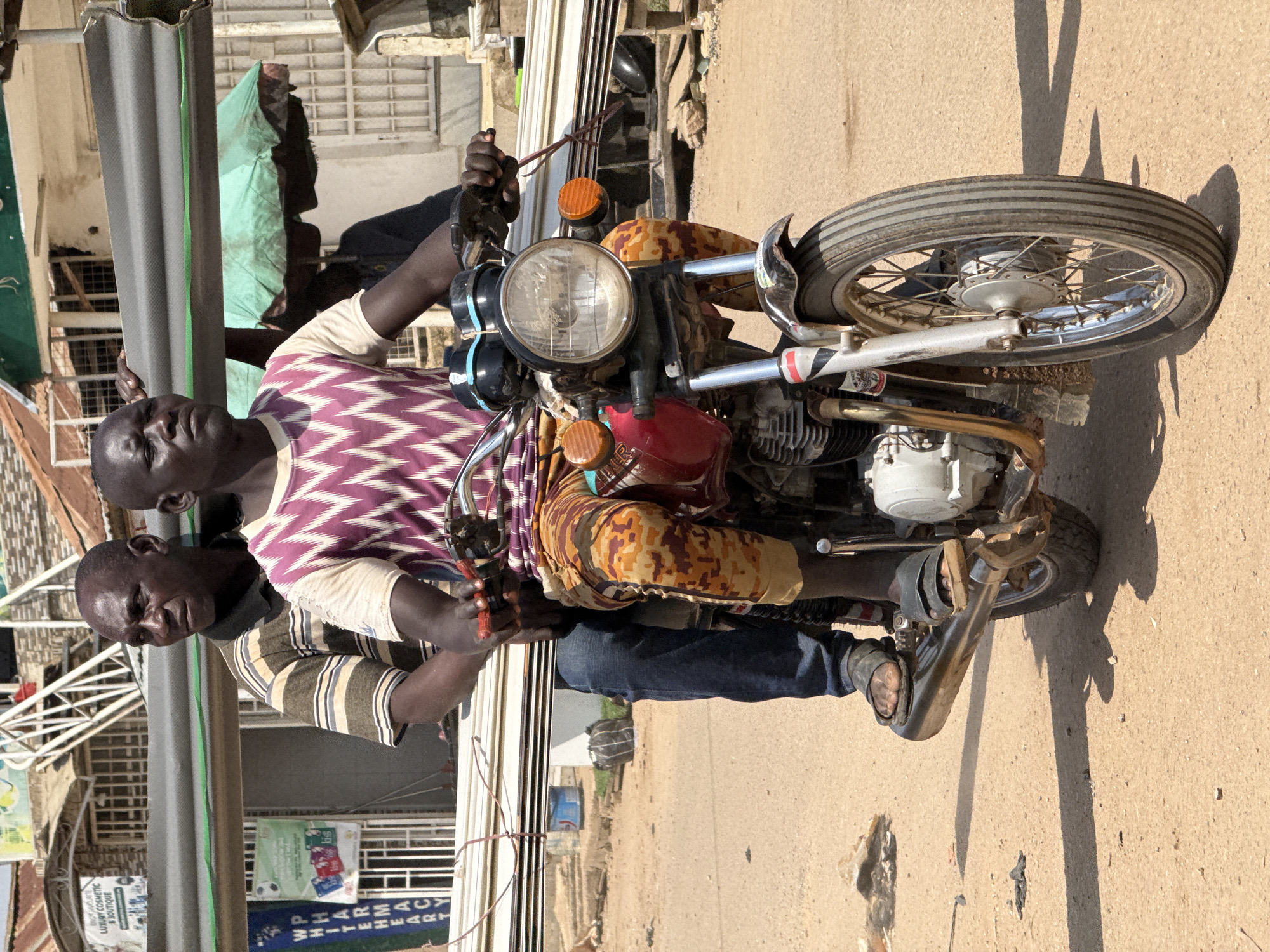 Two men are riding a motorcycle on a dirt road in Abuja, Nigeria. The man in the front is driving the motorcycle and is wearing a purple and white patterned shirt and camouflage-patterned pants. The man behind him is wearing a striped shirt and blue jeans. They are carrying long, thin objects, possibly building materials, on the motorcycle. The background shows buildings and shops, suggesting an urban or semi-urban environment. The lighting indicates it is daytime, with strong sunlight casting shadows.
