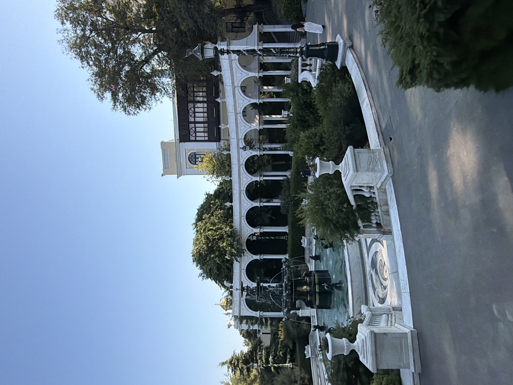 The image shows a view of a fountain and a white arched colonnade in a park on a clear day. The fountain is in the center of a pool with several decorative statues. The colonnade, with its classical columns and arches, forms a semi-circular backdrop to the fountain area. Behind the colonnade, a multi-story building with a light-colored facade and dark-paneled windows is visible. Lush green trees and bushes are abundant throughout the scene, creating a serene park-like atmosphere. A lamppost stands to the right of the colonnade, and a person in a white dress is walking away from the camera on a paved path. The paving in front of the fountain features a decorative mosaic pattern. The overall scene depicts a well-maintained outdoor public space with architectural and natural elements. The location is identified as the Fountain Square in Sabail Raion, Azerbaijan.