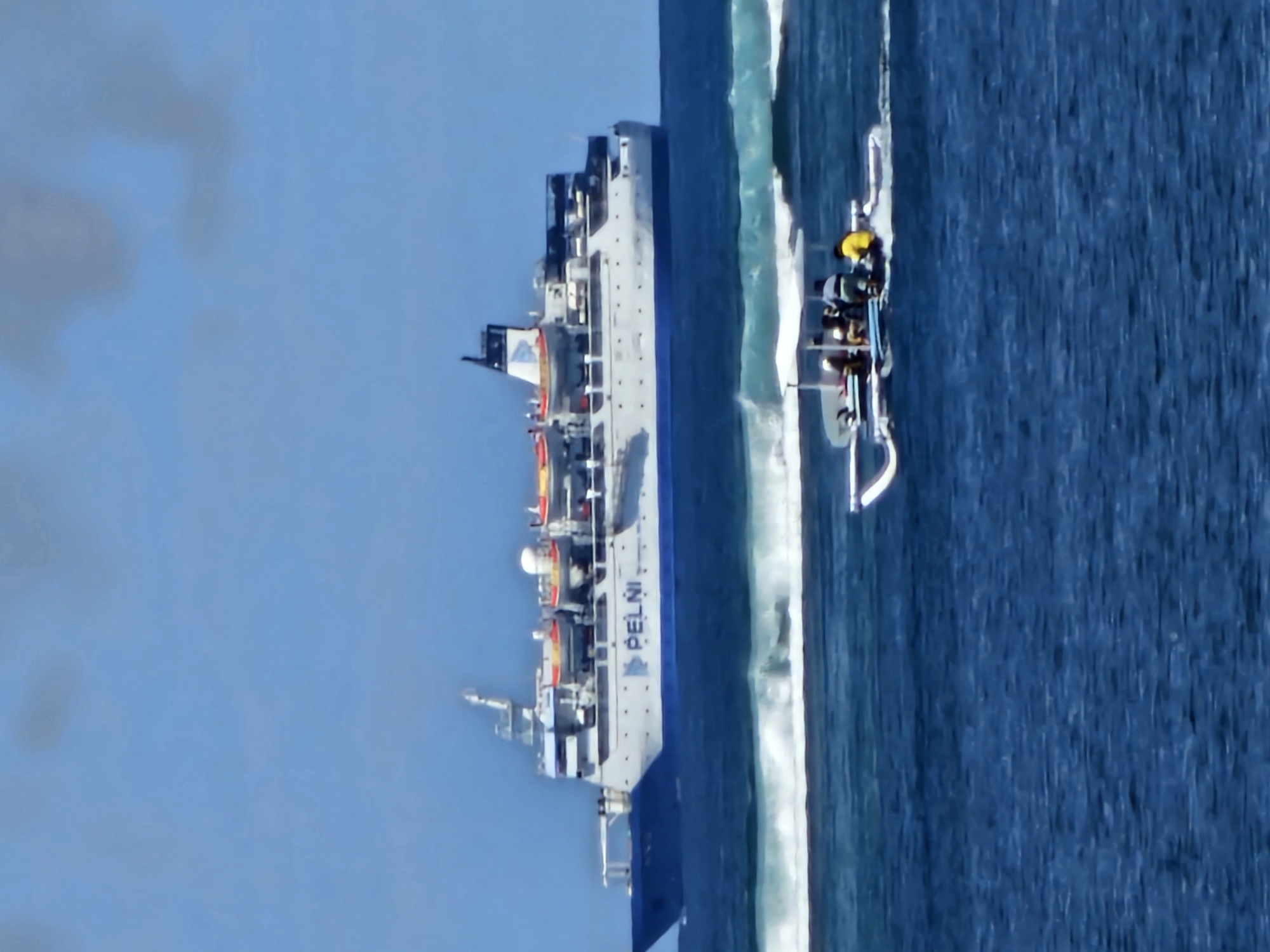 A large white ship with a blue hull sails on a vast blue ocean under a clear sky. The ship, labeled "PELNI," has several lifeboats and a tall structure at the back. In the foreground, a smaller white boat with a white canopy carries a group of people. The boat is moving, leaving a wake in the choppy blue water. Waves are breaking in the background, suggesting a shallow reef or coastline. The scene depicts maritime activity with a large passenger vessel and a smaller boat on a sunny day. The location is the waters near Denpasar, Indonesia.