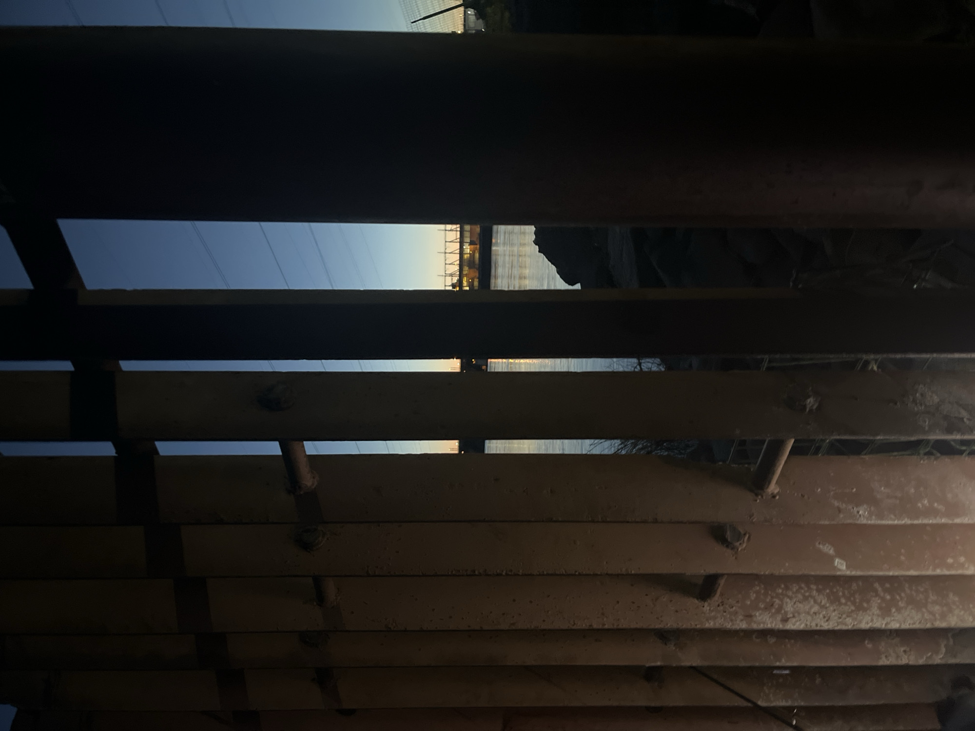 The image shows a view through vertical metal bars, resembling a fence or gate, at dusk. The bars are reddish-brown and appear weathered. Through the gaps between the bars, a body of water, likely a river, is visible, reflecting the twilight sky. Across the water, there is a distant structure with lights, possibly industrial or a bridge. Power lines can be seen strung across the sky. The foreground is dark and shadowed by the bars. The location is identified as being in the city of Chaikovsky, Russia.