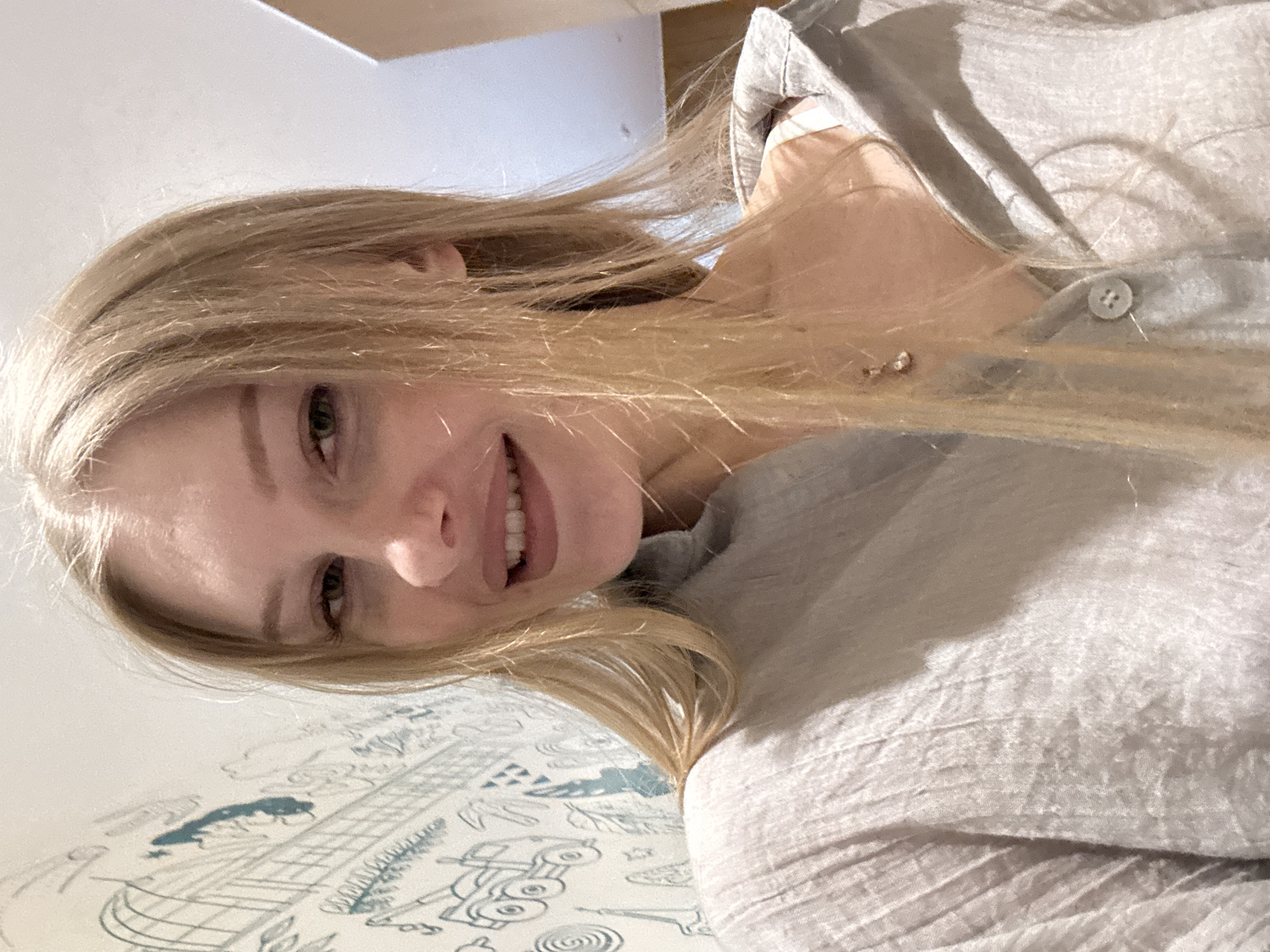A young woman with long, blonde hair is taking a selfie. She is wearing a light grey, textured shirt with a collar and a button. She has a subtle smile and her hair is falling over her shoulders and partially across her face. In the background, there is a white wall with a blue drawing of various objects, including what appear to be vehicles and buildings. To the right of the woman, a portion of a wooden structure is visible. The lighting suggests an indoor environment. The image does not contain any information that would allow for its specific location within the city of Perm, Russia.