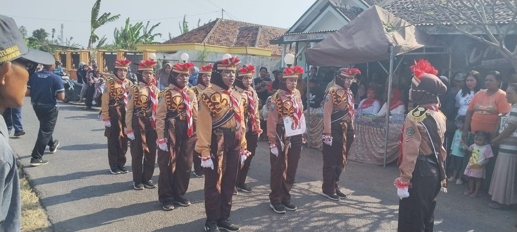In Yosowilangun Kidul, Indonesia, a group of young individuals dressed in scout uniforms are participating in a parade. The uniforms consist of tan shirts with brown collars and shoulders adorned with gold trim and badges, brown pants, and a red and white diagonally striped scarf. They are also wearing red and white feathered headwear. The scouts are marching in formation on a paved road, with spectators lining the sides. Behind the scouts, there are houses with tiled roofs, a tent set up, and various vehicles including motorcycles. The atmosphere appears to be festive, with people gathered to watch the event. The lighting suggests it is daytime, likely morning or afternoon, under a clear sky.