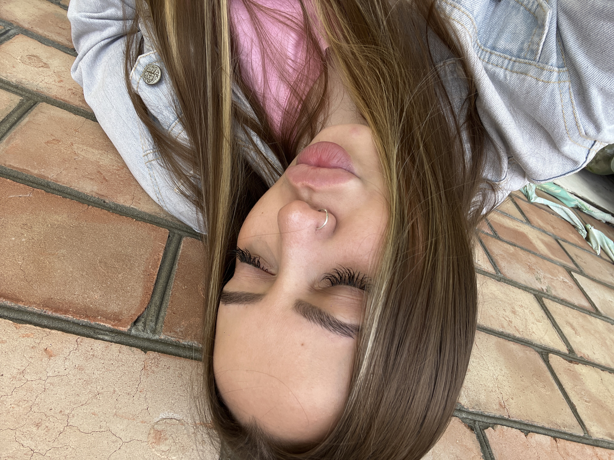A young woman with fair skin, brown hair with blonde highlights, closed eyes, and a nose ring is lying on a brick surface. She is wearing a pink shirt and a light denim jacket. The image appears to be taken from above, with the camera rotated. The location cannot be determined from the image.