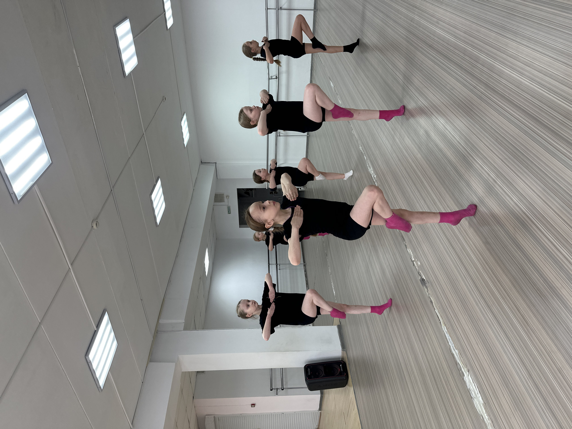 Six young girls are depicted performing a group exercise in what appears to be a dance studio. They are lying on their backs on a light-colored, wood-patterned floor, with their knees bent, feet flat on the ground, and arms crossed over their chests. All the girls are dressed in matching black short-sleeved tops and black shorts. Most are wearing vibrant pink socks, though one has black socks and another sports white socks.
The setting is an indoor studio featuring white walls and a white ceiling, which is fitted with numerous rectangular fluorescent light panels providing bright illumination. A significant portion of one wall is covered by large mirrors, reflecting the girls and the interior of the room, creating an illusion of greater depth. A silver ballet barre is visible running along the base of the mirrored wall. On the floor in the foreground, a black portable speaker, bearing the "SONY" brand, is positioned. A small green sign with white text, likely "ВЫХОД" (Exit), is visible in the reflection on a distant wall. The atmosphere suggests a focused practice or warm-up session. This event takes place in Votkinsk, Russia.