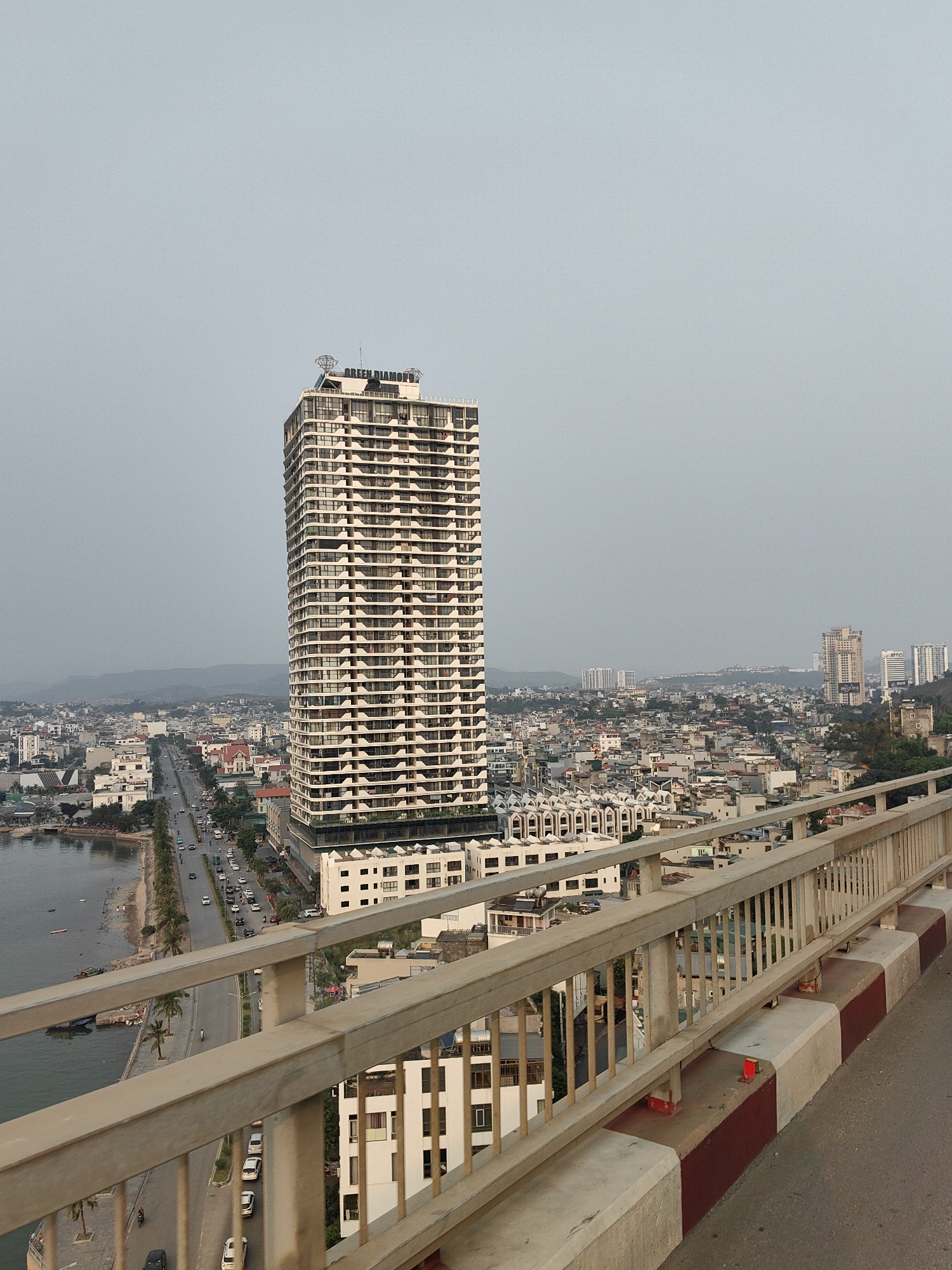 The image shows a tall, light-colored building with numerous balconies, labeled "GREEN DIAMOND" on top, in the Hong Gai Ward of Vietnam. The building is seen from an elevated viewpoint, likely a bridge, with a road running along the coastline to the left. Cars and other buildings are visible in the cityscape, which extends into the distance under a hazy sky. A bridge railing is visible in the foreground, with white and red markings along the curb.