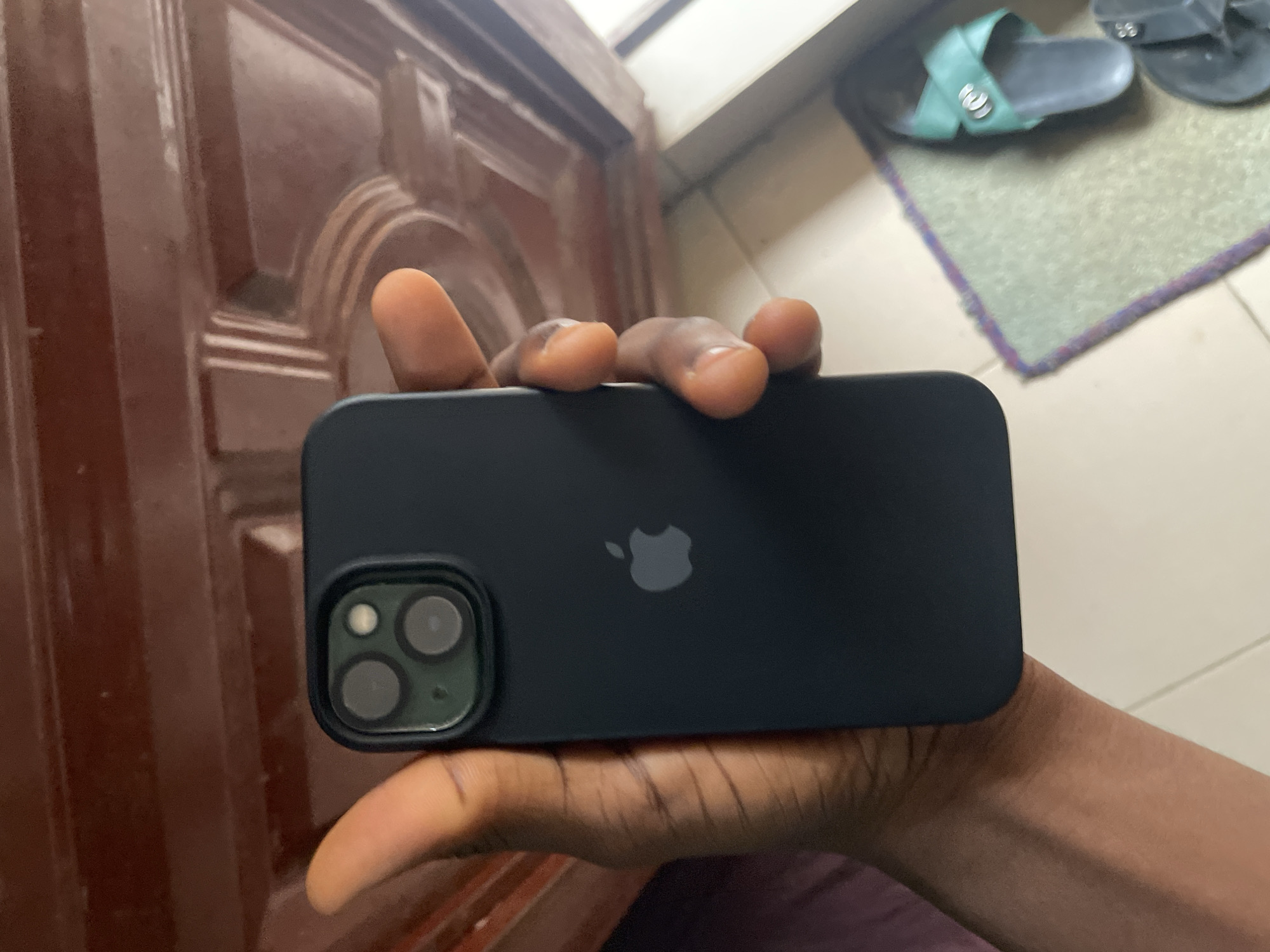 In Safari, Nigeria, a person's hand holds a dark-colored iPhone 13 Pro. The phone is positioned vertically, with the back facing the viewer. The camera module, located in the top left corner of the phone's back, contains three circular lenses arranged diagonally. Below the camera module, the iconic Apple logo is visible. The person's hand is positioned to grip the side of the phone, with fingers curving around the back. In the background, a wooden door with intricate carvings is partially visible. Below the door, a light-colored tiled floor is seen, with a textured rug and a pair of green flip-flops placed on it.