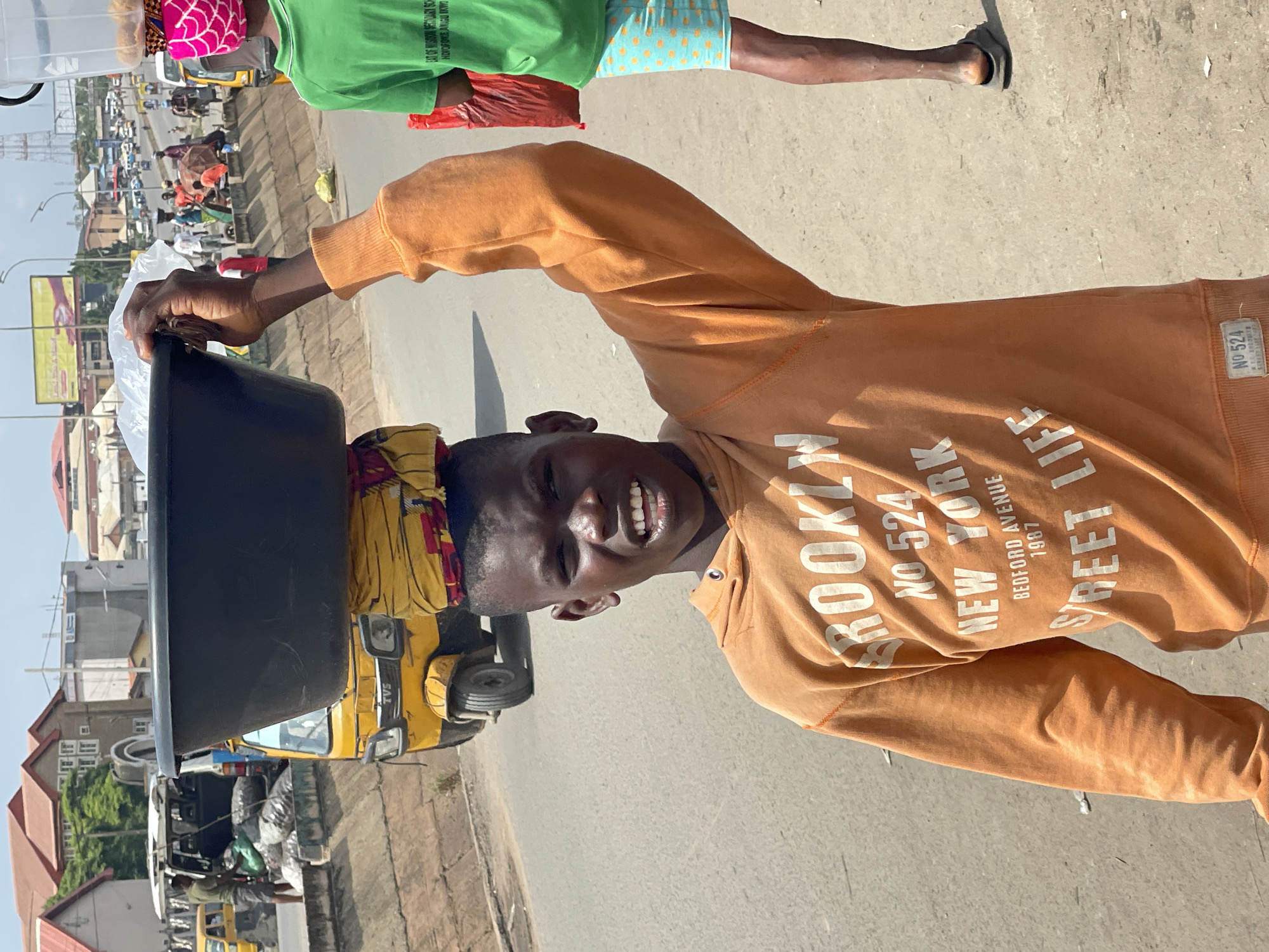 A young boy, appearing to be a vendor, balances a large black tub on his head, secured by a yellow and red patterned cloth. He is wearing an orange hooded sweatshirt with white lettering that reads "BROOKLYN NO 524 NEW YORK BEDFORD AVENUE 1987 STREET LIFE." He is standing on a paved road or sidewalk, with other people and vehicles in the background, suggesting an urban environment.
In the background, a yellow motorized tricycle (keke napep) is visible, as is a white minivan loaded with sacks. To the right of the boy, another person wearing a green t-shirt and patterned shorts is partially visible. The surroundings indicate a busy commercial area, likely a street market or roadside in Onitsha, Nigeria, known for its bustling trade. The presence of multiple vendors and vehicles suggests a typical day in this Nigerian city. The lighting indicates it is daytime, likely under a clear or partly cloudy sky.