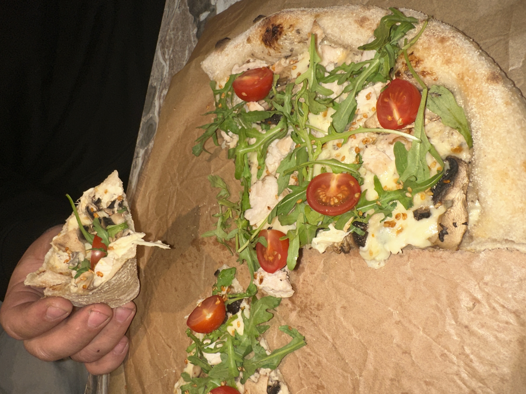 A close-up shot shows a partially eaten pizza on brown parchment paper. A hand holds a single slice, pulled away from the main pizza. The pizza appears to be topped with cheese, sliced cherry tomatoes, arugula, mushrooms, and what appears to be chicken. The crust is thick and slightly charred. The background includes a marble-like surface and a dark-colored garment. The photo's composition focuses primarily on the pizza and the hand holding the slice. It is impossible to verify the location is Chaikovsky, Russia, from the given visual data.