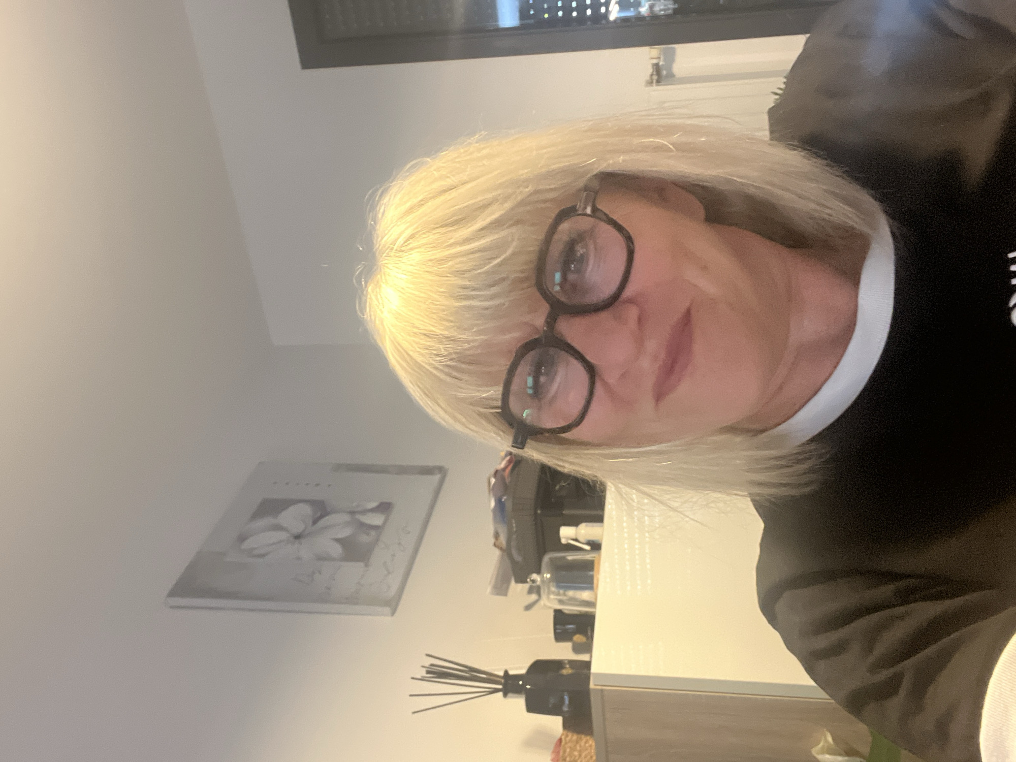The image shows a medium shot of a woman with blonde hair, wearing glasses and a black t-shirt with a white collar. She is looking at the camera and smiling slightly. Behind her, there is a white shelf with various items, including a black vase with reeds, a small glass dome, and a white spray bottle. A framed picture of a flower is hanging on the wall above the shelf. The scene appears to be indoors, possibly in a home or office setting, in Chamboeuf, France.
