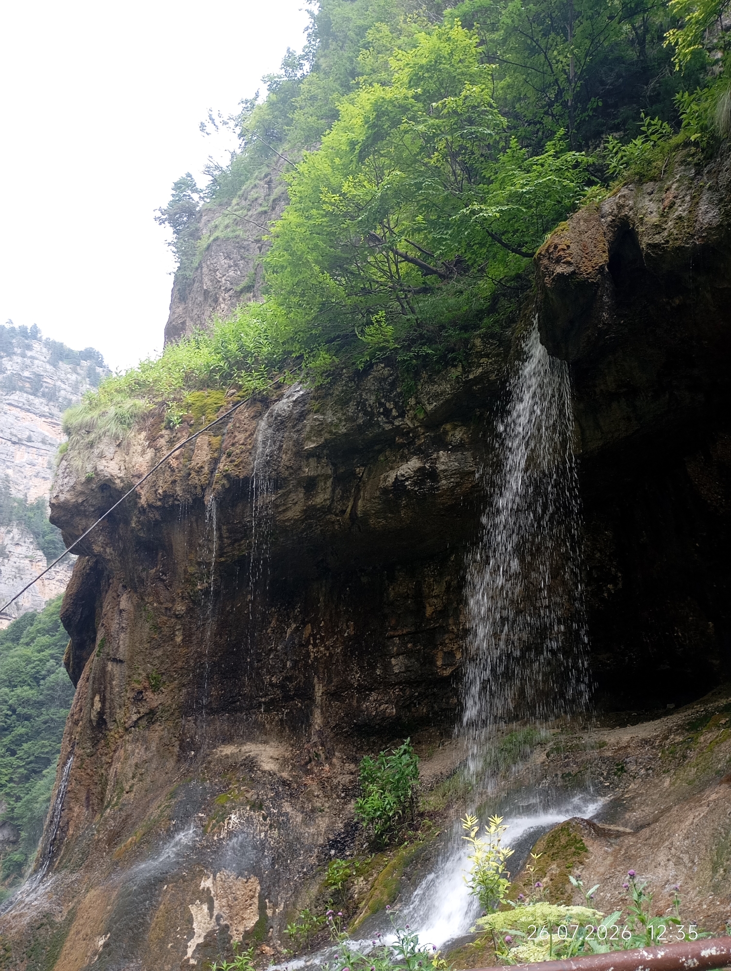 The image depicts a scenic view of a waterfall cascading down a rocky cliff face in Khushto-Syrt, Russia. Lush green vegetation, including trees and moss, grows along the top and sides of the cliff. The waterfall is the focal point, with water falling in a curtain-like fashion into a stream or pool at the base. A metal cable is visible running diagonally across the left side of the cliff. The environment appears natural and somewhat rugged, with the overcast sky suggesting a potentially cool or humid day. The timestamp "26.07.2026 12:35" is visible in the bottom right corner of the image, indicating the date and time the photograph was taken. There are no people or animals visible in the image.