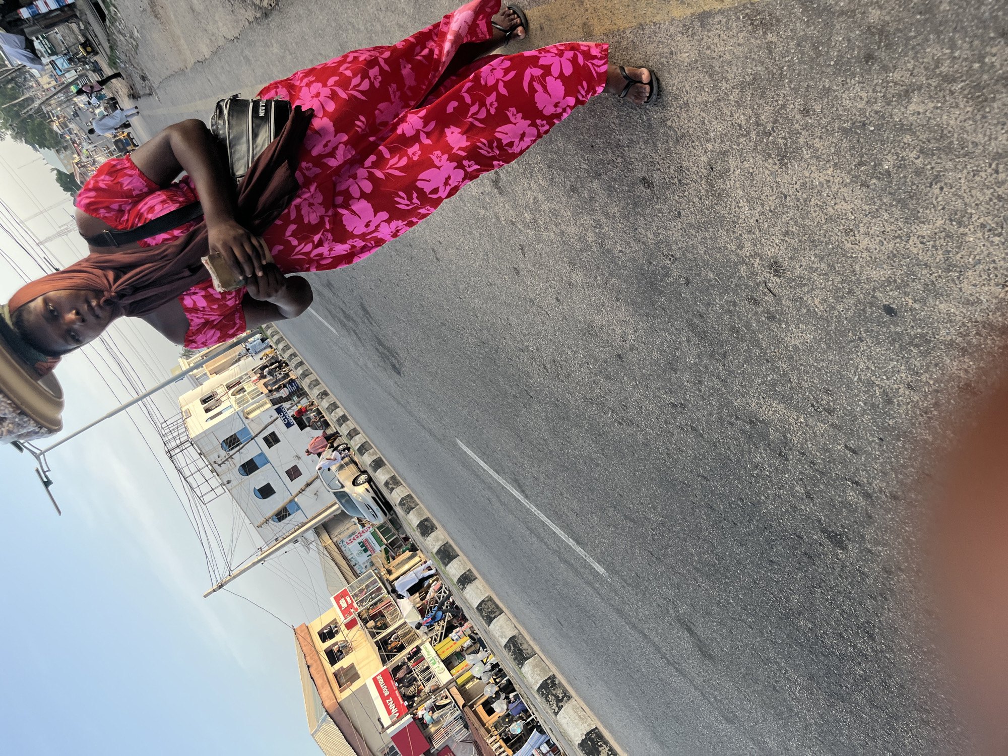 In an outdoor, urban environment, a woman of African descent is captured mid-stride on a paved street. She is wearing a vibrant red dress adorned with a pink floral pattern and a brown headscarf. A dark-colored crossbody bag with the word "NEW YORK" printed on it is slung over her shoulder. Her gaze is directed forward, and she holds what appears to be a mobile phone in her right hand. A stack of items is balanced on her head, suggesting she may be a vendor or carrying goods. The background reveals a street scene with various buildings, including shops and commercial establishments, and a few other individuals walking on the sidewalk. The sky above is clear and displays a soft blue hue. The image is shot from a slightly low angle, emphasizing the woman and the surrounding environment.