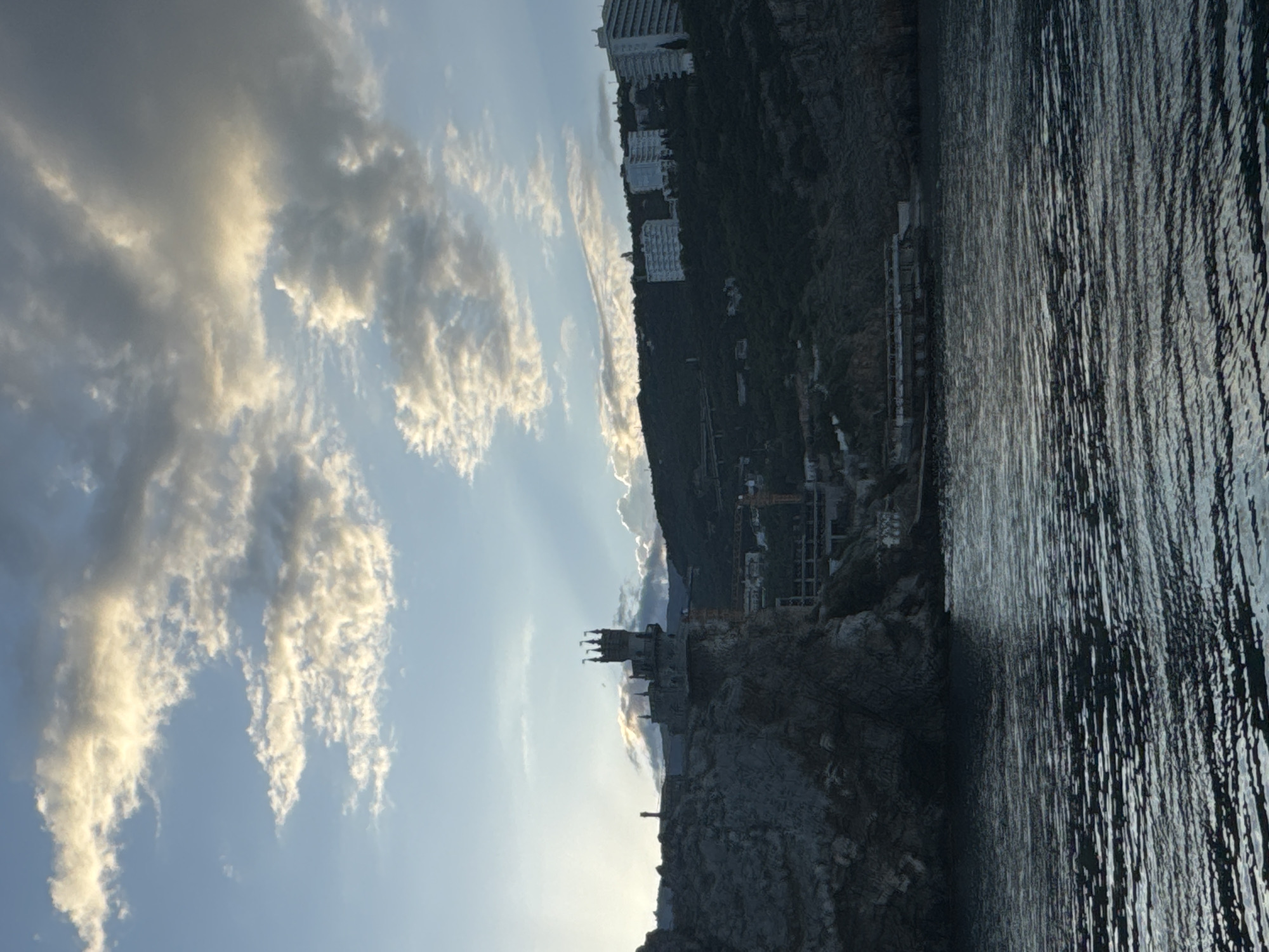 The image depicts the Swallow's Nest castle, a historical landmark located in Гаспринский поселковый совет, Ukraine. The castle is perched on a rocky cliff overlooking the Black Sea, with the water body occupying the right side of the frame. The horizon is partially obscured by the cliff on which the castle sits and the mountainous terrain to the left. The sky, occupying the top half of the image, is filled with clouds, and the lighting suggests either sunrise or sunset. The architecture of the castle is visible against the sky. The image appears to be taken at an angle, possibly from a boat or another vantage point at sea level.