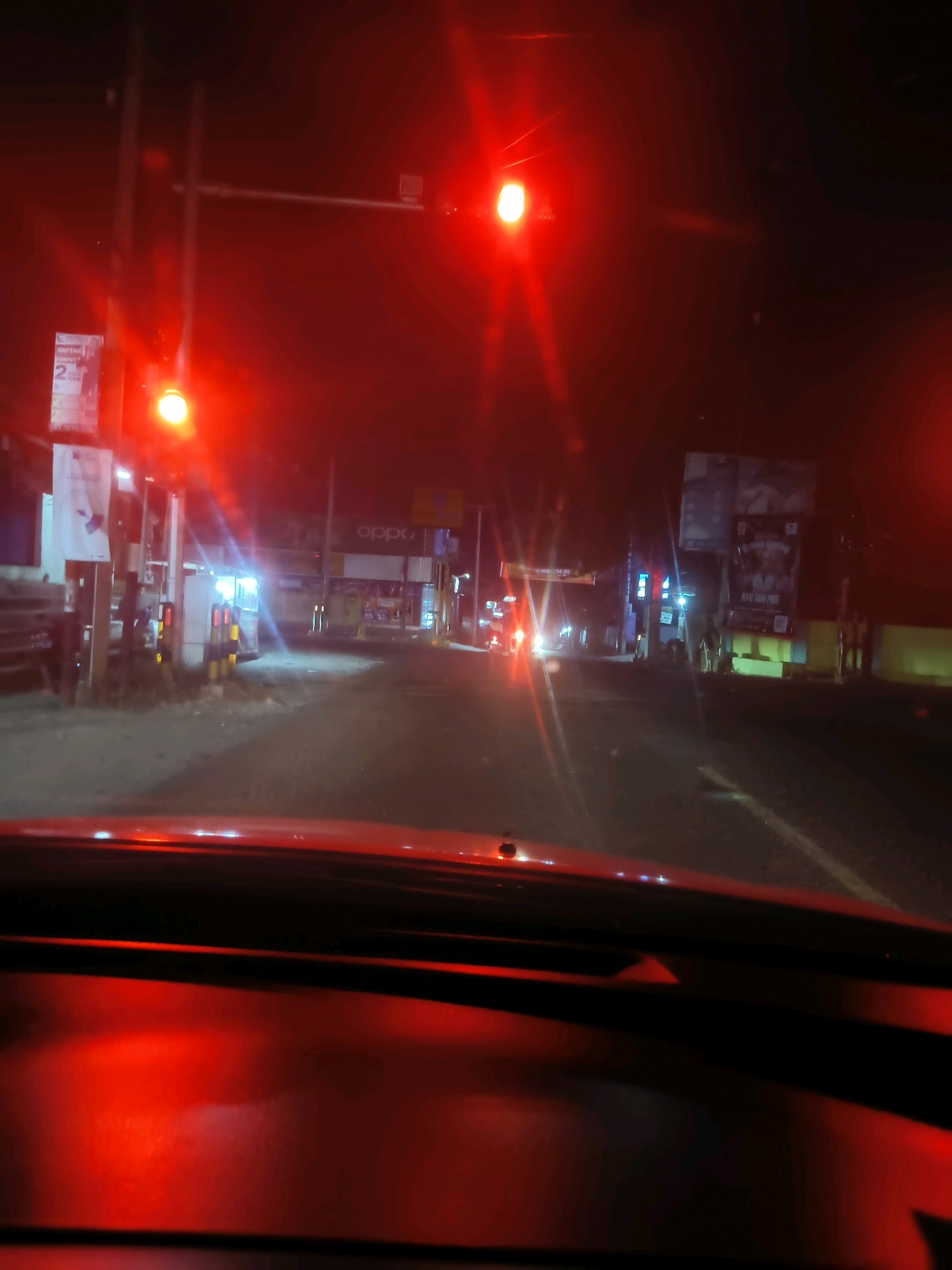 The image depicts a street scene at night in Serang, Indonesia, likely at an intersection controlled by a traffic light. The dominant color is red due to the illuminated traffic signal, casting a warm glow over the surroundings. The perspective is from inside a vehicle, as suggested by the view of the dashboard and windshield.
In the foreground, the red dashboard of a car is visible, reflecting the light from the traffic signal. The road ahead is dimly lit, with a lane marking visible.
The midground shows a street with traffic lights. The main traffic light, which is red, is prominently displayed on a pole. There are also some signs and advertisements visible on poles along the side of the road. One sign appears to be for "OPPO". Other vehicles or people are not clearly discernible in the midground, though the presence of streetlights suggests a populated area.
The background is darker and more obscured by the nighttime conditions and the red light. However, it appears to be a typical urban street with buildings and possibly more signage. There are some distant lights that suggest further activity in the city.
The overall impression is a snapshot of a moment at a traffic light in a city at night, characterized by the pervasive red light and the limited visibility typical of such conditions.
