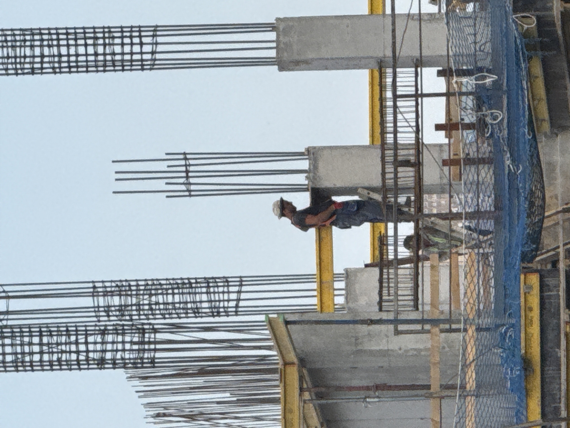 The image shows a construction site in Tbilisi, Georgia. A construction worker wearing a white hard hat, dark shirt, and blue pants is visible. The worker is positioned on a scaffold, leaning over to work on a rebar structure. The scene depicts an active construction project, with concrete beams and numerous rebar elements forming the framework of a building. Yellow scaffolding poles and a blue safety net are also prominent features. The overall impression is one of ongoing construction work, likely during daylight hours given the clear sky. No text is visible in the image.