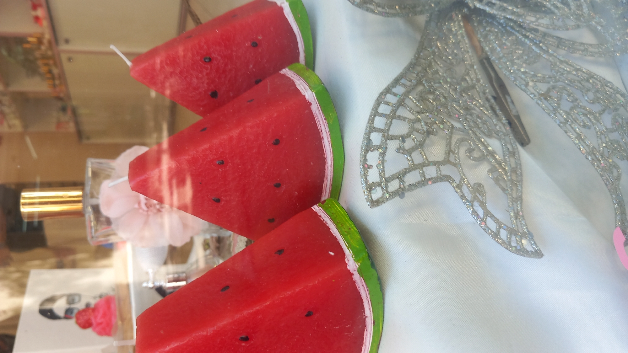 The image depicts a close-up view of a display window, likely in a shop located in the city of Kos, Greece. The foreground is dominated by three wedge-shaped candles designed to resemble slices of watermelon. These candles are a vibrant red with black dots representing seeds, and a green rind at the bottom, with a thin white band separating the red from the green. To the right of the candles, a decorative silver bow, adorned with glitter, is visible.
In the background, a perfume bottle with a gold cap stands prominently behind the candles. To the left of the perfume bottle, a framed artwork features a stylized illustration of a woman wearing sunglasses, with a pink, strawberry-shaped object in front of her. The entire scene is viewed through a glass pane, indicated by reflections on the surface. The lighting suggests an indoor setting, possibly a boutique or a gift shop.