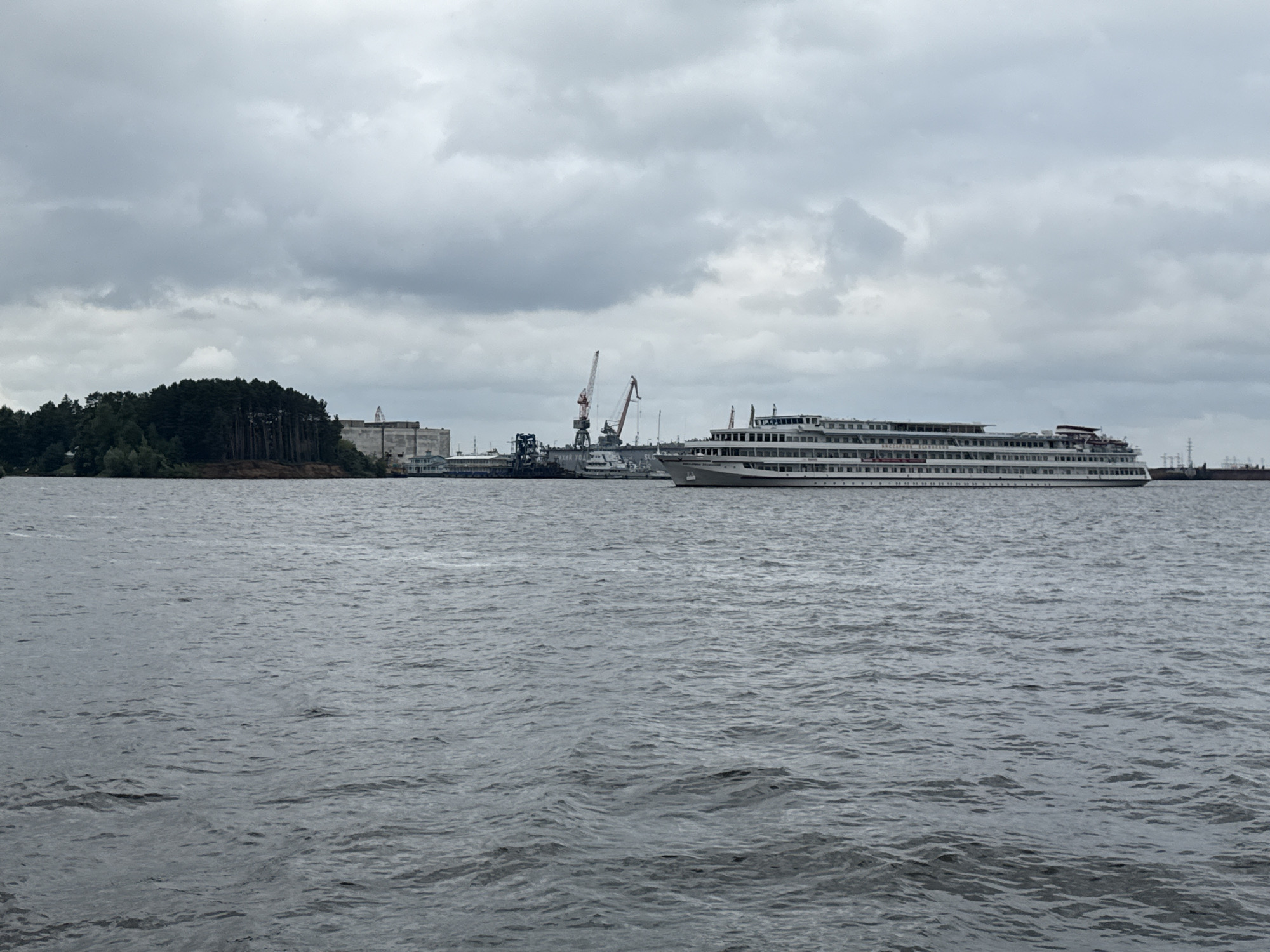 The image depicts a wide, calm river with choppy water in the foreground. On the far bank, a long, multi-deck passenger ship is docked. Behind the ship, a cityscape is visible, characterized by industrial buildings and port cranes. A forested hill with some cleared areas is situated to the left of the industrial zone. The sky is overcast with heavy clouds. The scene is likely the Kama River adjacent to the city of Chaikovsky, Russia, known for its shipbuilding and port facilities. There are no people or animals clearly visible in the image.