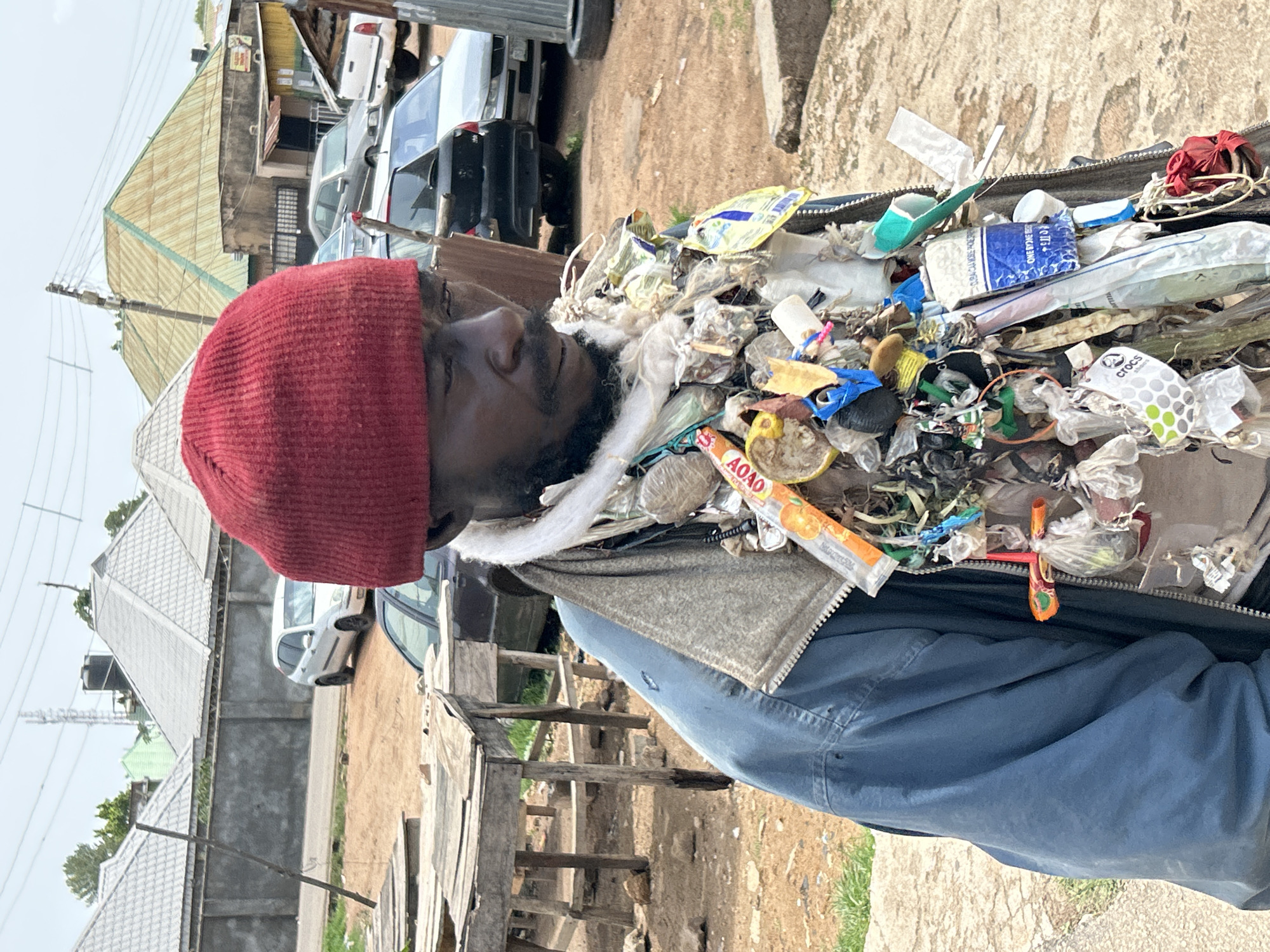 The image shows a man wearing a red beanie and a blue jacket, adorned with various items including plastic bags, wrappers, and what appear to be personal trinkets. He is standing outdoors, with a dirt road and some buildings in the background. The environment suggests an urban or semi-urban setting, possibly in Jos, Nigeria, based on the architectural styles of the buildings and the presence of vehicles. The man's attire and the items attached to his clothing could indicate a personal expression or a form of street artistry. The scene appears to be captured during daylight hours.
