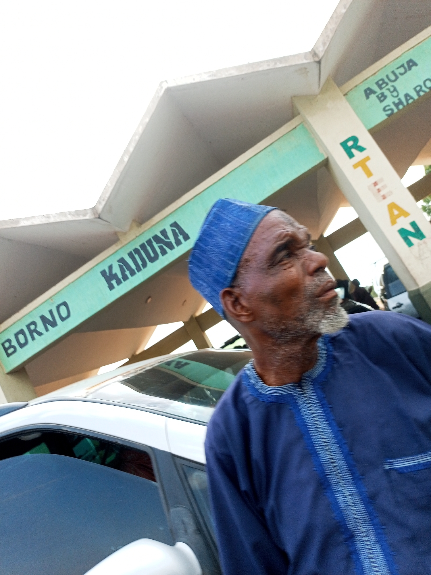 An outdoor medium shot captures a man with a grey beard and blue cap, wearing a blue tunic with intricate trim, looking to his right. Behind him, a building structure with a light-colored roof and green trim is visible. The green trim displays black text that reads "BORNO KADUNA" on one section and "ABUJA BY SHARON" on another, interspersed with vertical text on a white beam reading "RTAN". A white car is parked in front of the building, with its reflective surface showing parts of the structure and sky. The lighting suggests an overcast day.
