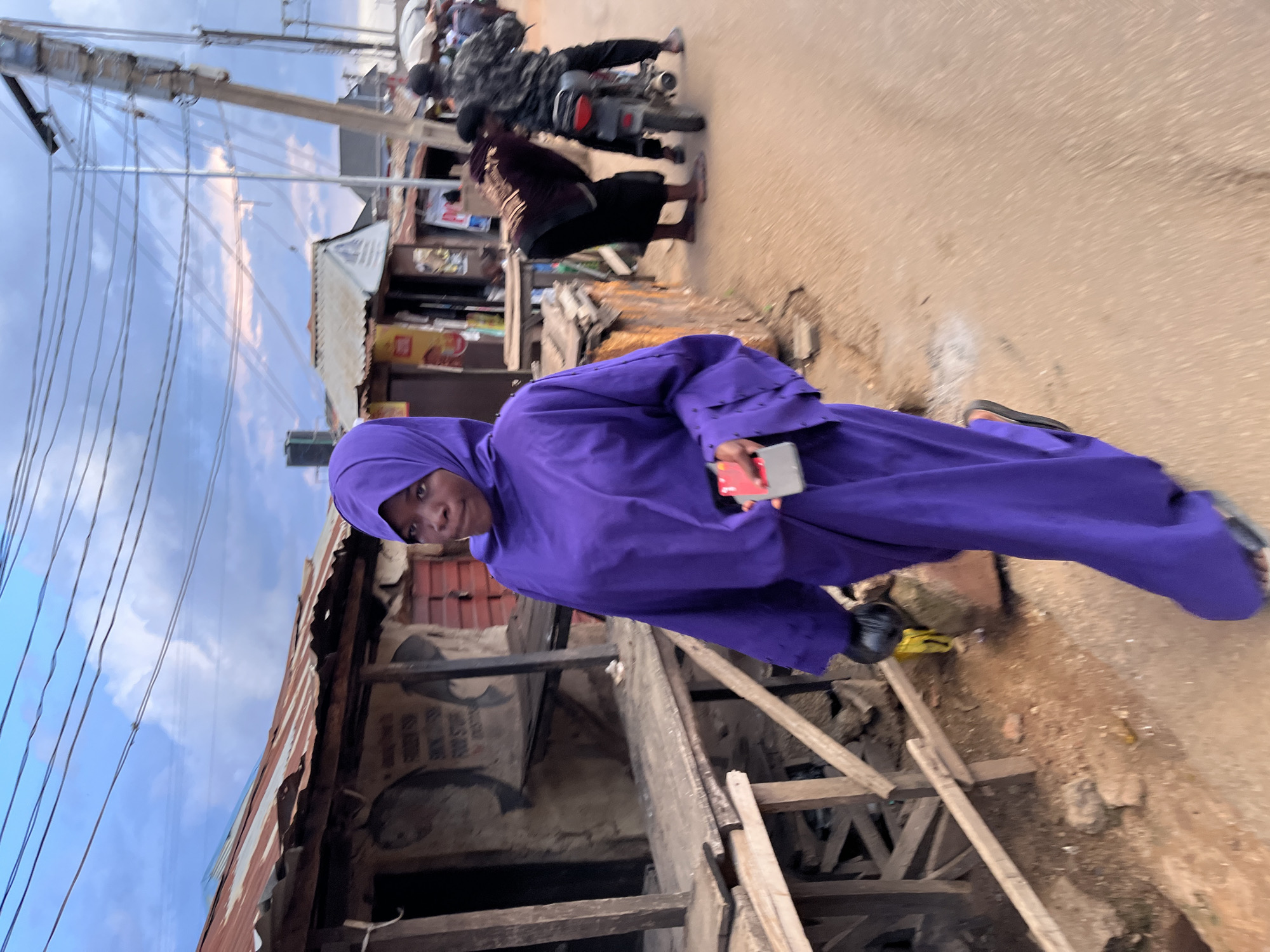 The image, rotated 90 degrees clockwise, depicts an outdoor street scene with a central figure and bustling background activity. A person, the primary subject, wears a vibrant, full-length purple garment, possibly a jilbab or abaya, with a matching head covering (hijab). They look towards the camera with a slight smile and hold a small red object, resembling a smartphone or card, in their right hand.
The setting is an unpaved, light brown dirt road. To the left of the main subject, a structure with a corrugated iron roof and exposed wooden support beams suggests basic construction or a state of disrepair. A brown box or sign behind the person in purple bears faded text, partially legible as "SUDAN POU" and "NO. STORE."
In the background, other individuals are visible on the street. One person is riding a motorbike, indicating movement and traffic. Another person, dressed in darker clothing, stands nearby. Overhead, a complex network of utility poles and numerous electrical wires crisscrosses against a cloudy, bright sky, suggesting daytime. The scene conveys a sense of daily life in an active, possibly urban or semi-urban environment.