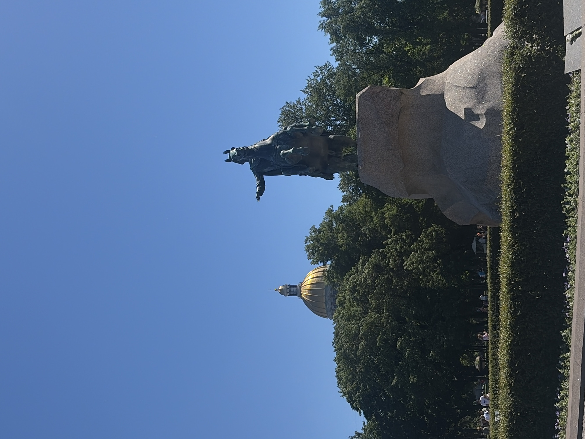 The image depicts a sunny day in Saint Petersburg, Russia, featuring the Bronze Horseman monument and a glimpse of St. Isaac's Cathedral in the background. The Bronze Horseman, a statue of Peter the Great on horseback, is positioned prominently in the foreground, dramatically posed on a large, rough-hewn stone base. The equestrian statue is dark bronze in color and points its right arm forward. Behind the monument, lush green trees form a dense backdrop, partially obscuring the golden dome and upper section of St. Isaac's Cathedral, which is visible to the left. The sky is a clear, bright blue, indicating fair weather. In the middle ground, a meticulously manicured dark green hedge runs horizontally across the scene, separating the monument area from a park-like setting. Several people are visible in the park, appearing as small figures walking or standing amidst the greenery, engaged in leisurely activities. The overall atmosphere is serene and suggests a popular tourist or public space.