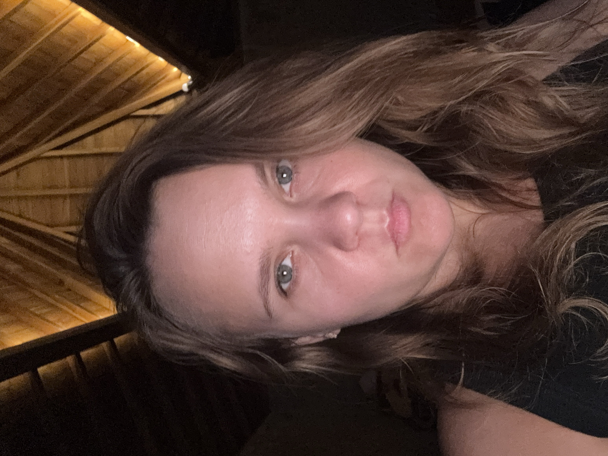 A close-up, forward-facing, indoor selfie shows a Caucasian woman with medium-brown, wavy hair and light-colored eyes. She is wearing a dark-colored top. The background is dark with an overhead view of a wooden, sloped ceiling, possibly with lighting fixtures visible. The lighting is dim and warm, casting shadows on the subject and background. The location is unspecified within the city of Unknown, Unknown.