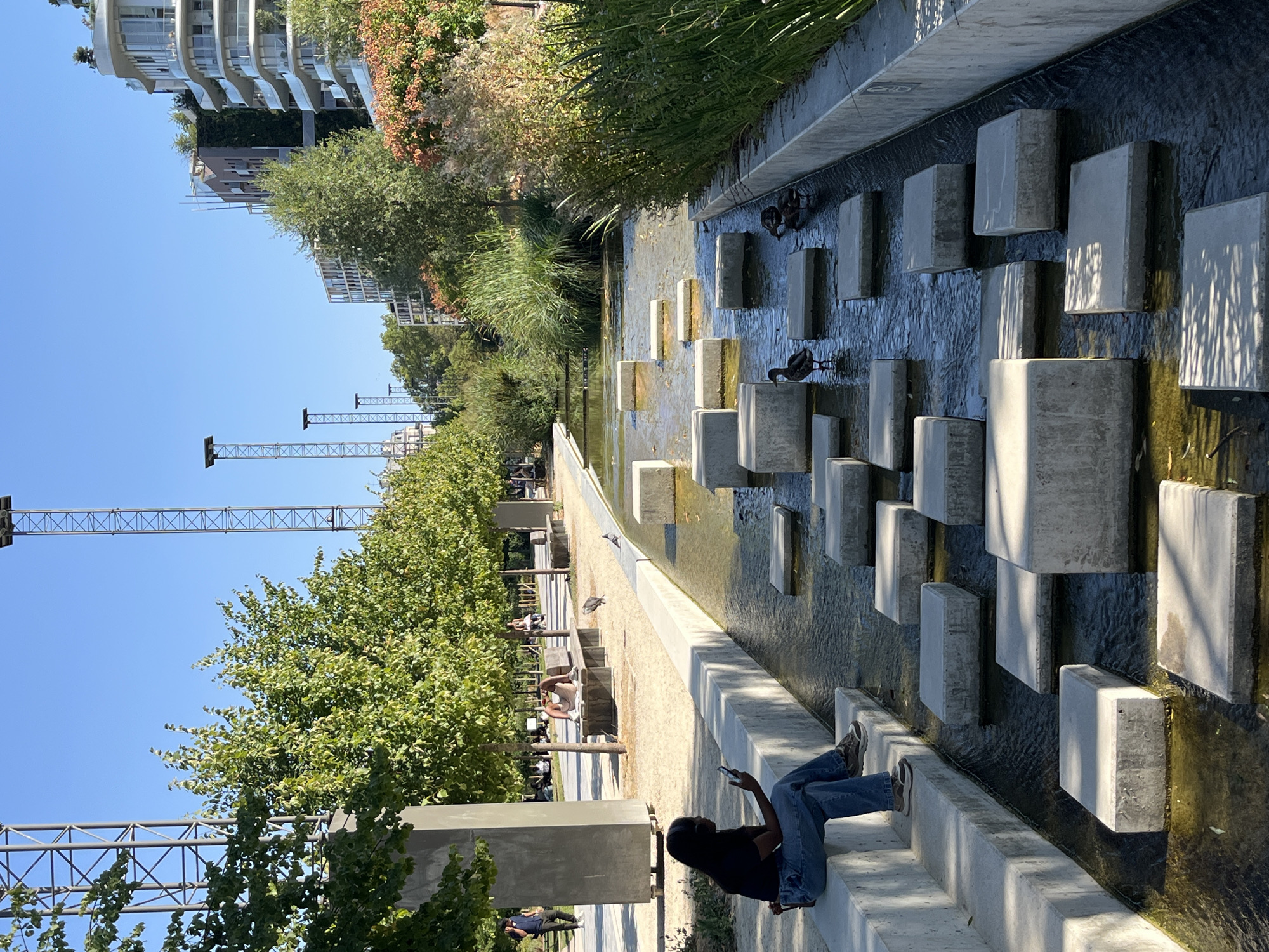 The image depicts a sunny day in a modern urban park, identified as Parc André-Citroën in Paris, France. A woman is seated on a concrete ledge, looking at her phone. The park features a shallow water channel with stepping stones, on which several ducks are visible. Tall metal towers with lights are present in the background, alongside contemporary buildings and lush green trees. Other people are scattered throughout the park, some sitting on benches or walking. The scene is characterized by clean lines, concrete structures, and a vibrant natural setting.