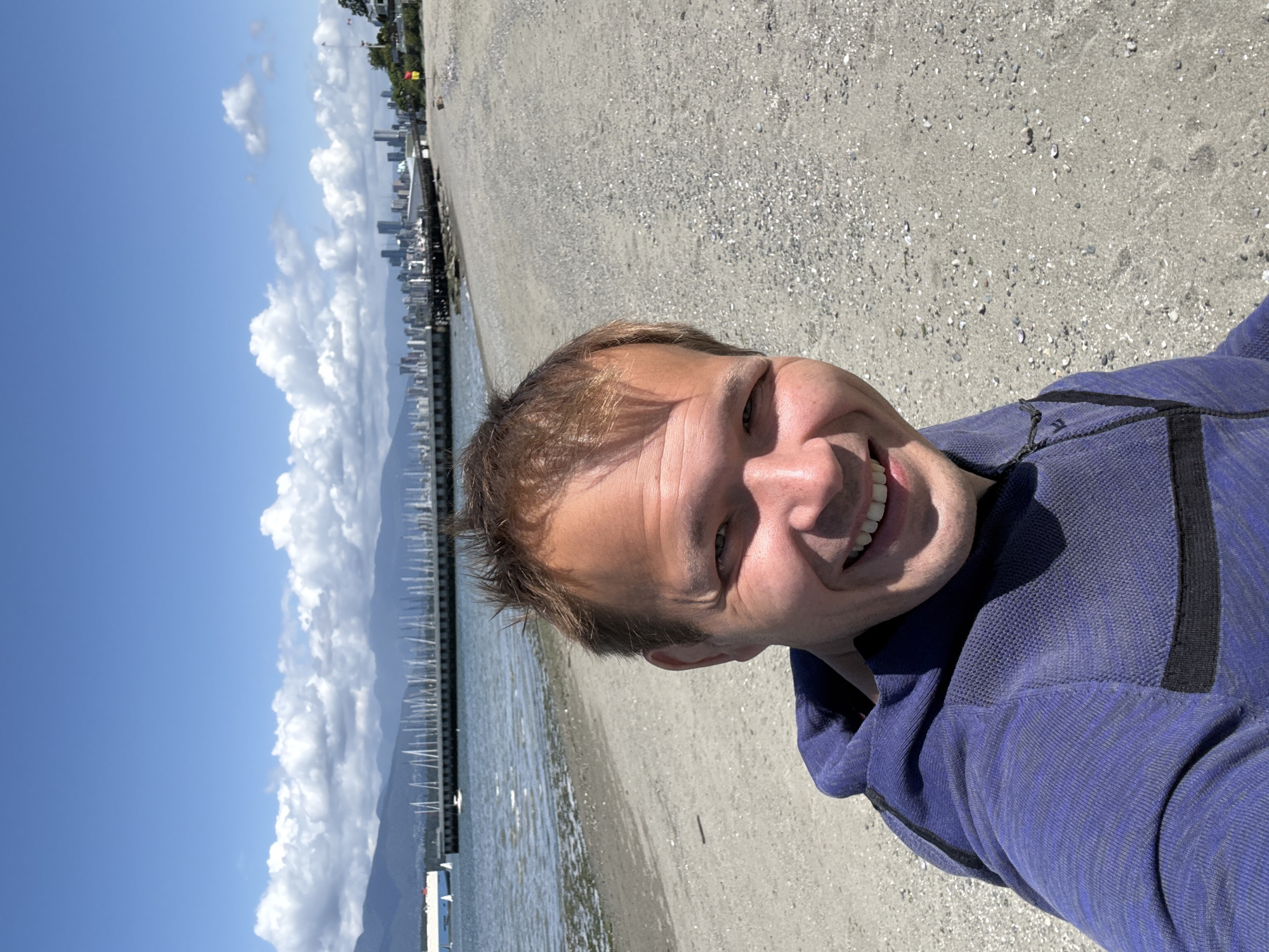 A selfie captured in Vancouver, Canada, shows a smiling man wearing a purple hooded sweatshirt. The backdrop reveals a beach alongside the water with a view of downtown Vancouver on the horizon. A pier with boats is visible on the water. The sky is blue with fluffy white clouds. The ground appears to be sand and gravel.