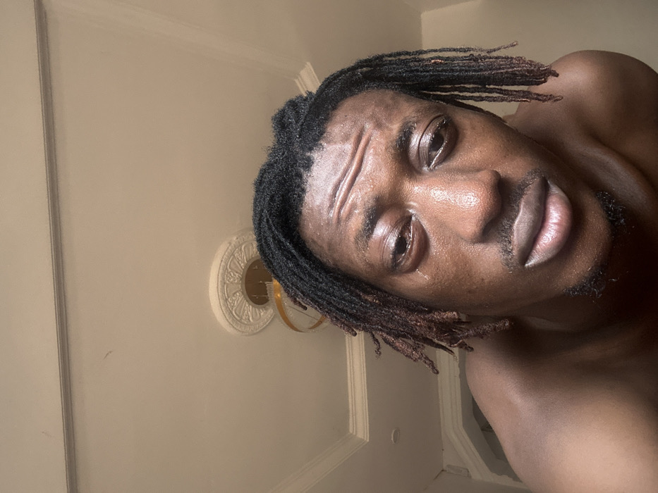 An indoor close-up photograph depicts a male subject of African descent, appearing to be an adult, from the chest up. The individual has dark dreadlocks with some reddish-brown coloration near the ends, a mustache, and a goatee. His facial skin, particularly the forehead and cheeks, appears moist and glistening. A distinct tear is visible near the inner corner of the subject's left eye, which, along with the right eye, appears somewhat heavy-lidded. Horizontal creases are present on his forehead. The subject is shirtless, with the upper portion of his right shoulder and chest visible. The background reveals a light-colored ceiling with a white decorative medallion surrounding a light fixture that features a golden-toned element and a clear shade. White crown molding is present along the top of the wall, and a light-colored architectural archway is partially visible on the far left. The scene is consistent with an interior room. The provided location of Egbe, Nigeria, cannot be independently verified through visual analysis of the image.