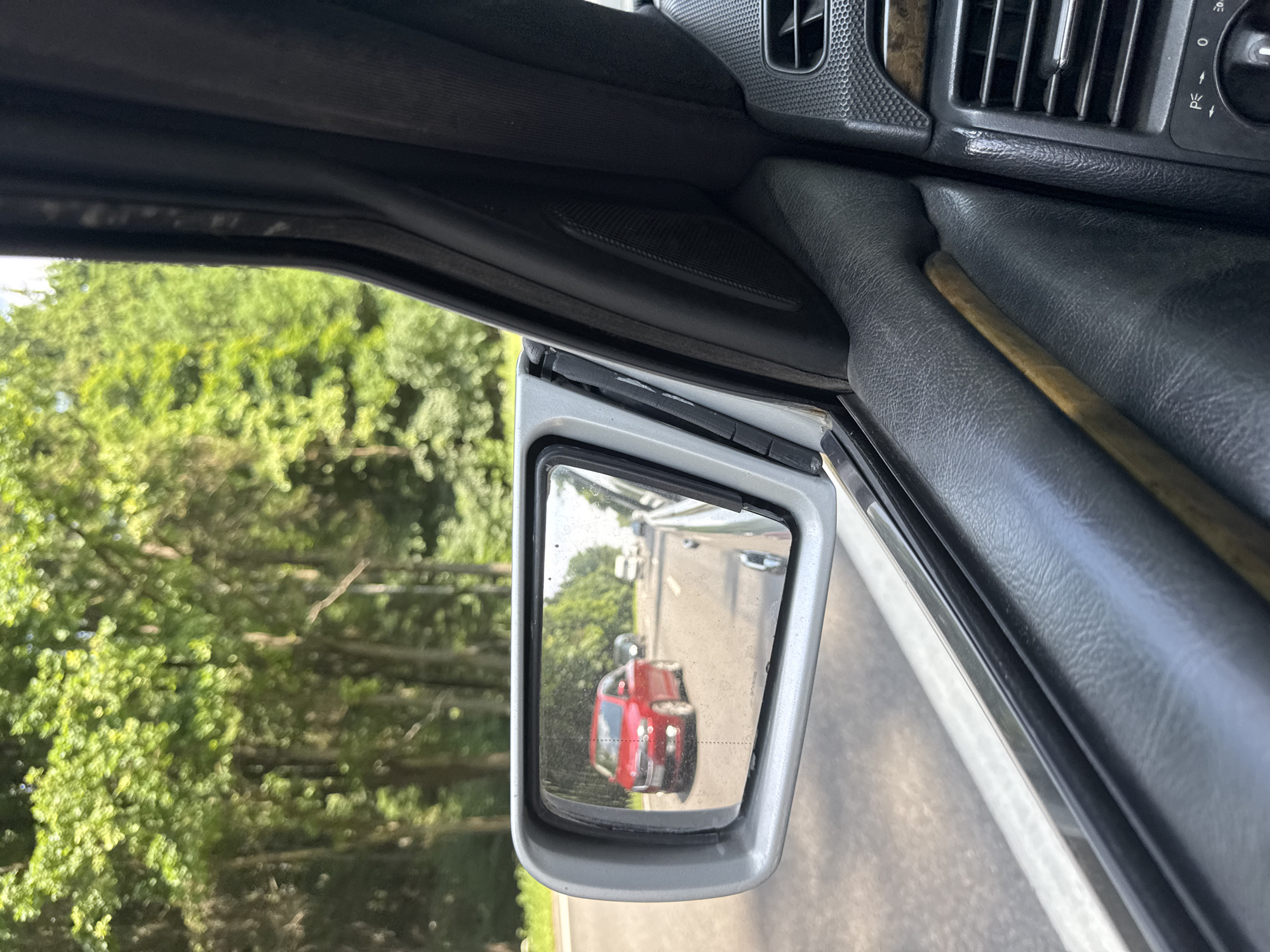 The image is taken from inside a car, looking out the passenger-side window and into the side-view mirror. The mirror reflects a red car driving on a road bordered by lush green trees. The road appears to be a highway or a well-maintained rural road. The interior of the car is visible, including part of the dashboard with ventilation grilles and a wooden trim. The scene suggests a car journey through a natural, wooded environment, likely during daylight hours. The location is identified as Schomyšlicki sel'ski sovet, Belarus.