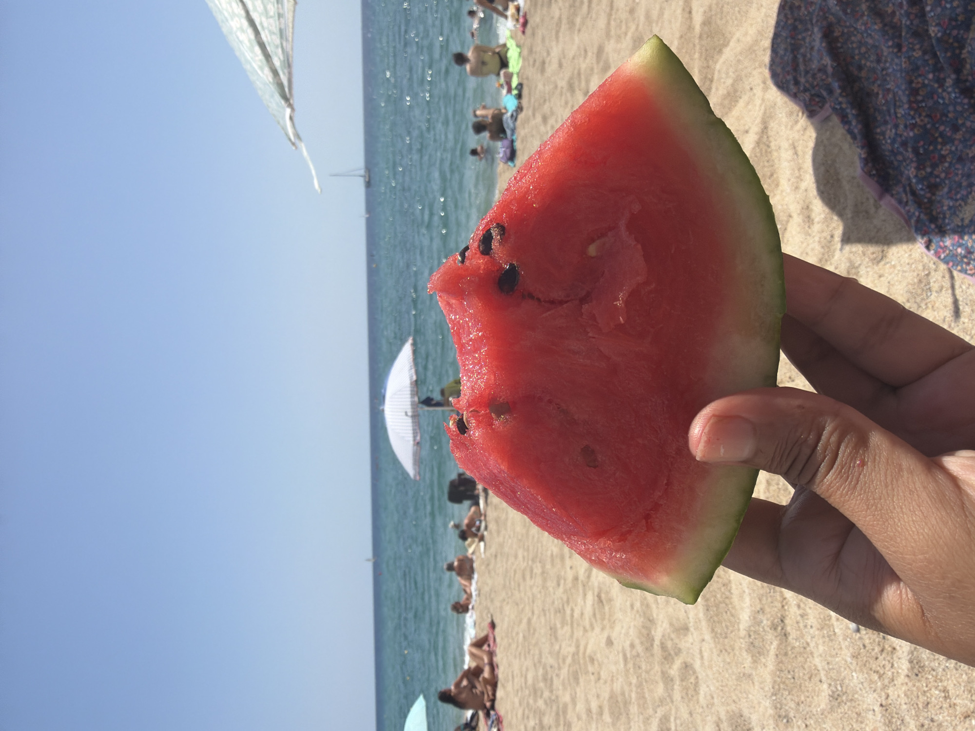 A close-up shot shows a person's hand holding a slice of watermelon at the beach. The watermelon is bright red with black seeds and a green rind. In the background, the sea is visible under a clear blue sky. Several beach umbrellas and people are scattered on the sand, some lounging and others in the water. The scene appears to be a sunny day at a beach in Badalona, Spain, with the Mediterranean Sea in the distance.