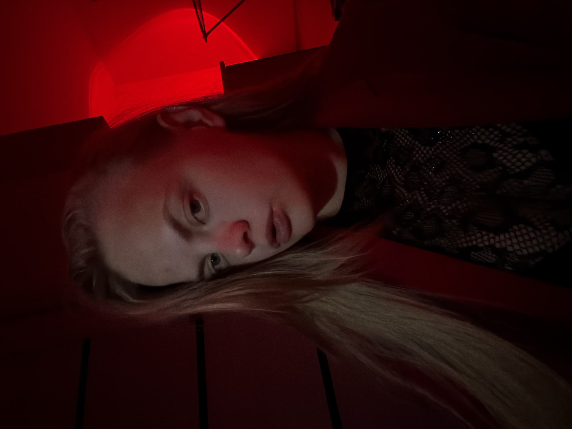 The image is a close-up, dimly lit selfie taken indoors, possibly at night, with a strong red lighting source. The subject is a young female with blonde hair, wearing a dark, lacy top and a red blazer. Her gaze is directed towards the camera with a neutral expression. The background is mostly obscured by shadow, but appears to be a wall with horizontal lines, and a blurred red circular light source on the right side. No specific location in Izhevsk, Russia, is identifiable from the image.