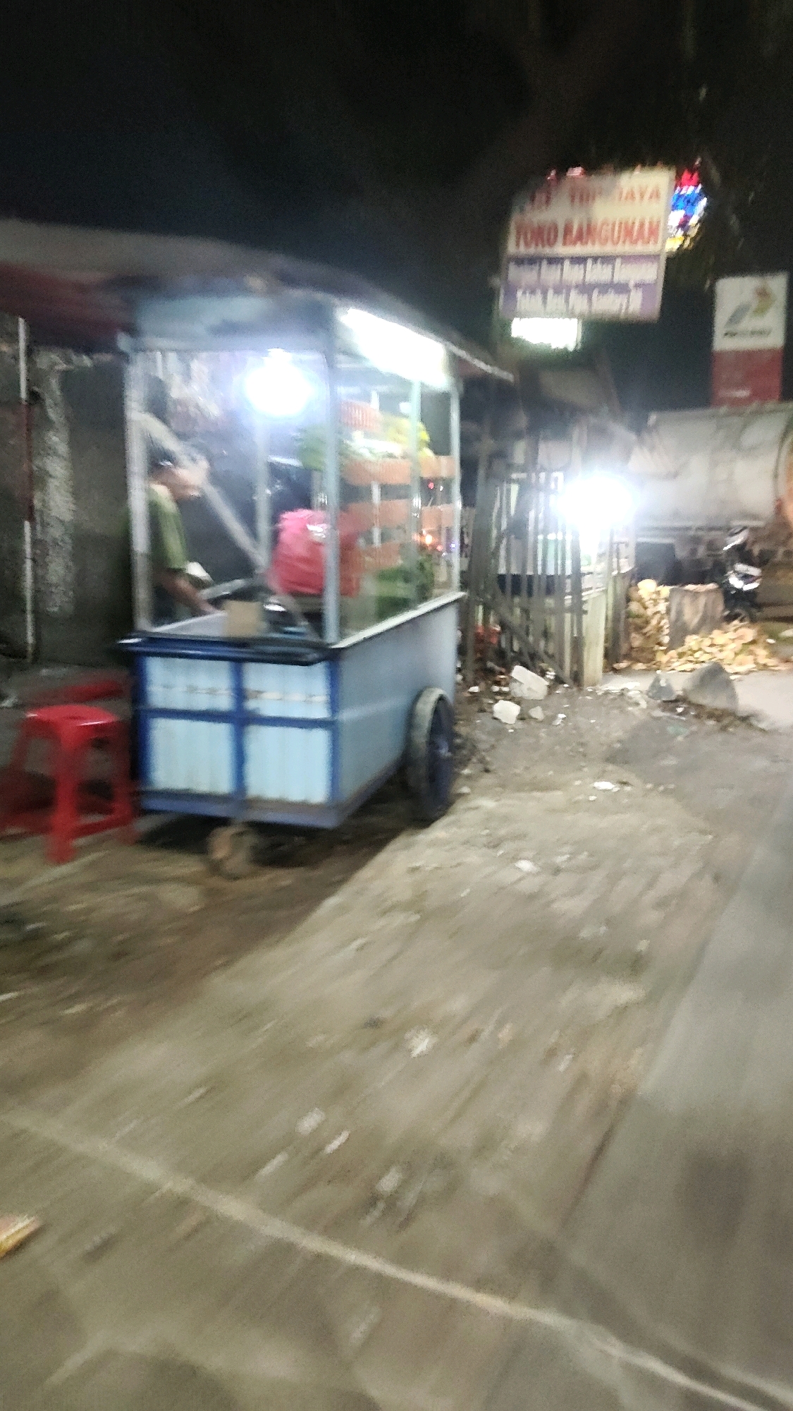 The image shows a blurred view of a street scene at night in Mekarmukti, Indonesia. A food cart, possibly selling juices or fruits, is illuminated by bright lights, displaying an assortment of items in baskets. A red stool is placed in front of the cart. In the background, a sign that reads "TOKO BANGUNAN" (Building Store) is visible, along with a portion of what appears to be a gas station. The ground is a dirt or gravel surface. The overall scene suggests a commercial area at night.
