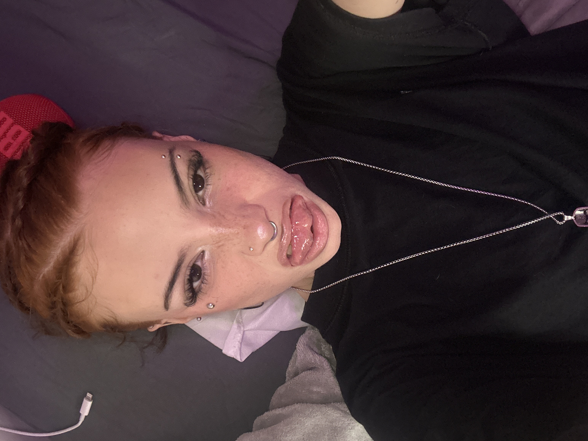 A young woman with auburn hair styled in braids is lying on a grey bed with a black top on, extending her tongue. She has piercings on her eyebrow, nose, and ear. A silver chain with a pendant hangs around her neck. A red shoe is in the background. A white charging cable is visible near the bottom left of the frame. The location is not identifiable within the city of Buenos Aires, Argentina based solely on the image provided.