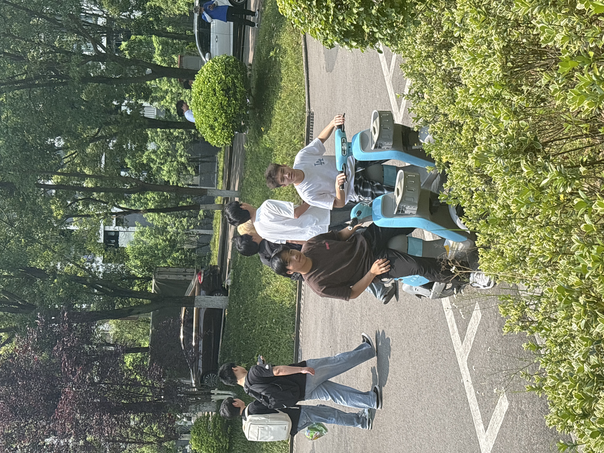 A group of people are gathered around a light blue tandem bicycle on a paved surface. One person in a brown shirt is on the front seat, while another in a white shirt is on the back. Two other people are walking on the road. The environment is lush with greenery, trees, and bushes. In the background, a car is parked, and buildings are visible. The location is likely in Jiangjin, China, based on the environment and people.