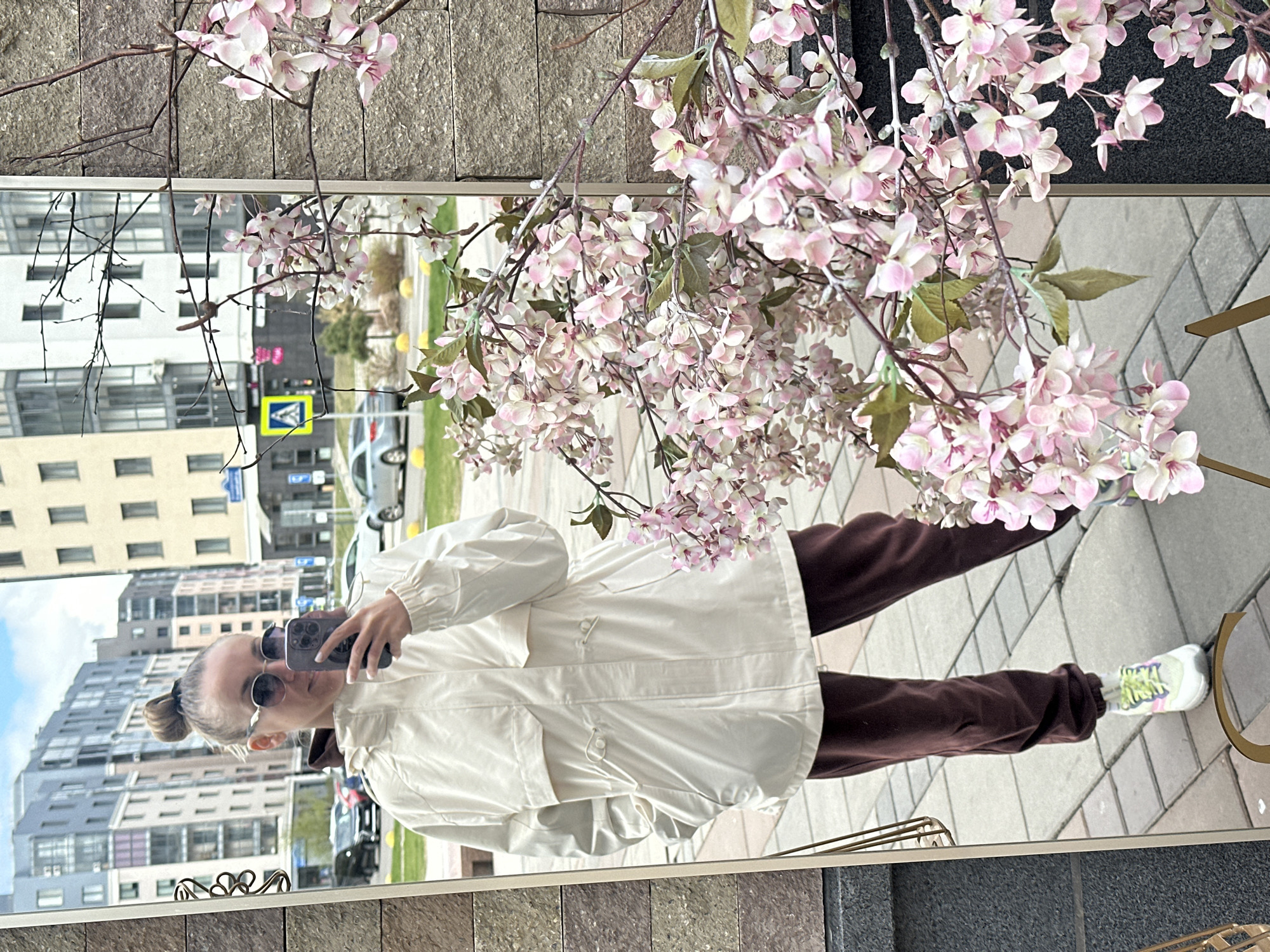 A woman in sunglasses, a light-colored jacket, and brown sweatpants is taking a mirror selfie. The mirror reflects an urban landscape, buildings, and street signs, consistent with a city. Pink flowering branches partially obscure the reflection. The location is Krasnoyarsk, Russia.