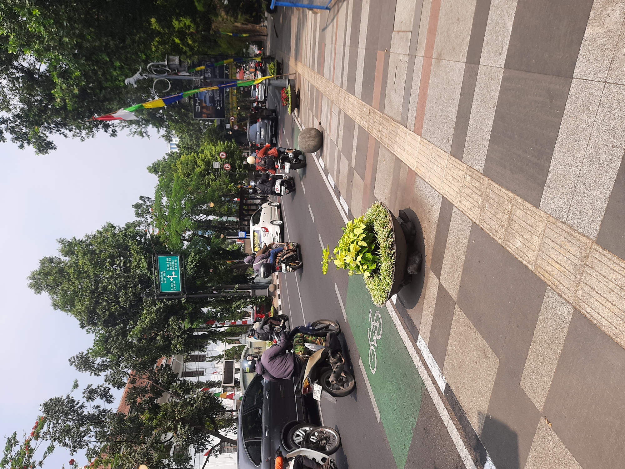 The image depicts a street scene in Bandung City, Indonesia, likely on a sunny day. The street is paved with asphalt and features a dedicated green lane for bicycles marked with a white bicycle symbol. On the right side of the street, a tiled sidewalk is visible, with textured tactile paving for visually impaired pedestrians. A large, round stone is placed on the sidewalk near a planter containing green leafy plants.
Vehicles are present on the road, including several motorcycles with riders wearing helmets and casual clothing, a dark-colored SUV, and white cars. Indonesian flags, red and white, are displayed on poles along the roadside. A green directional signpost indicates destinations such as "Jalan Ir. H. Juanda," "Tugu", "Alun Alun", "Pasar Baru", "Banda", "Husein Sastranegara", and "Lembang". The background shows buildings and lush green trees, suggesting an urban environment with ample vegetation. The overall atmosphere is one of moderate urban activity.
