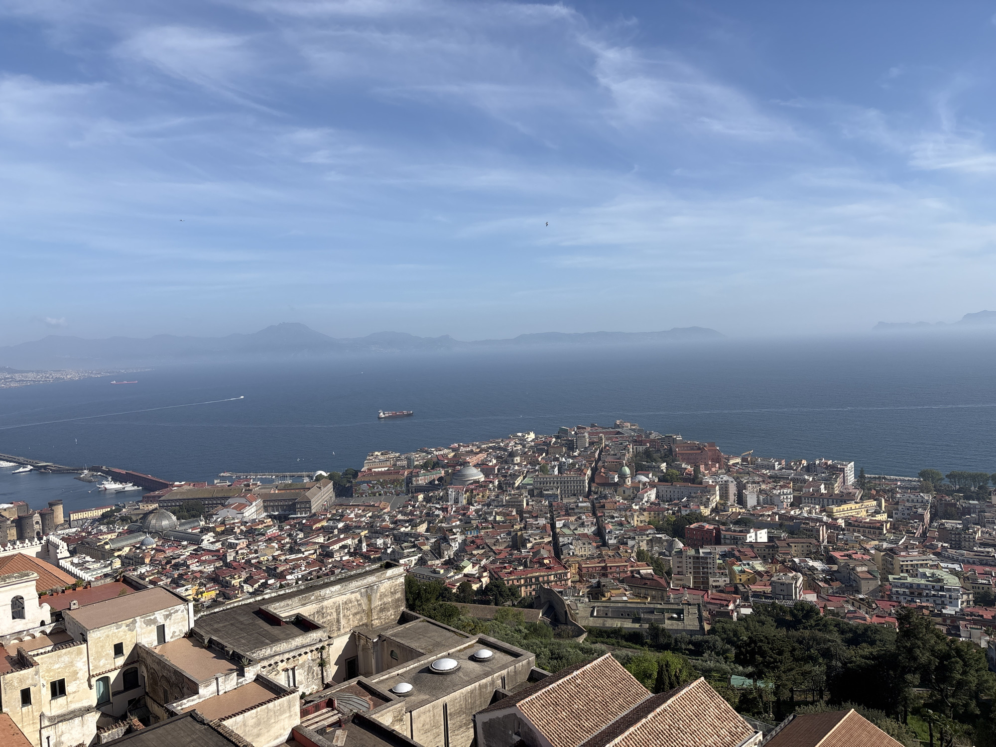The image is a high-angle, daytime view of the city of Naples, Italy, overlooking the Bay of Naples. The city is densely built, with a mix of residential and commercial buildings visible. Many of the buildings have red-tiled roofs. In the foreground, there are rooftops of buildings with various architectural features, including domes and arched windows. Further out, there is a coastline with harbors, and a mountainous region in the distance. The sky is blue with scattered clouds. The sea is calm with a few ships visible.
