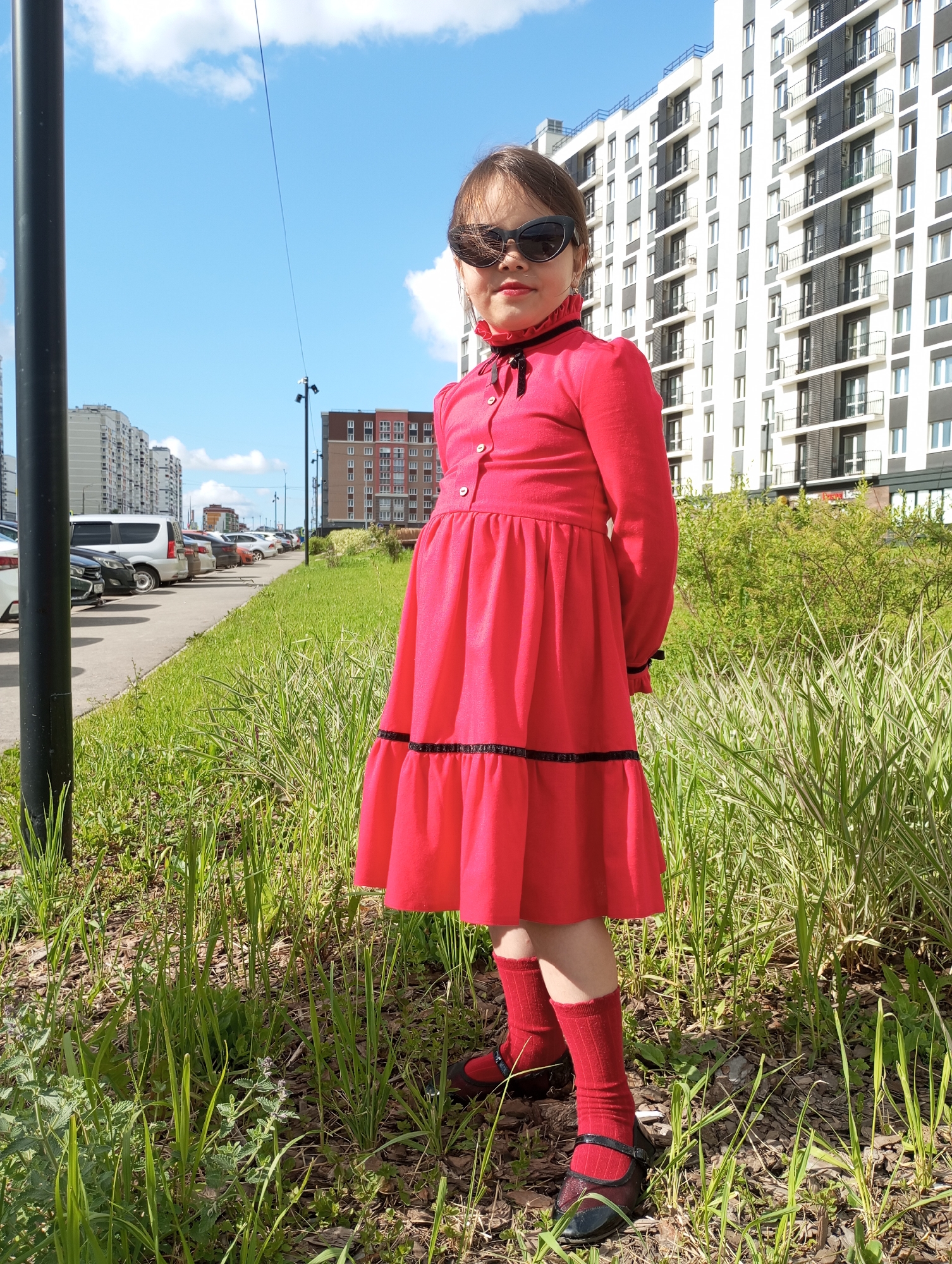 A young girl stands outdoors on a bright, sunny day in a residential area of Pervomaysky, Russia. She is centrally positioned, looking directly at the camera with a slight smile visible beneath her dark, cat-eye sunglasses. She wears a vibrant red long-sleeved dress featuring a ruffled high collar with a black ribbon bow, and black trim details on the skirt and cuffs. Her attire is completed with red socks and shiny dark Mary Jane style shoes.
The background presents a mix of urban and green elements. In the immediate foreground, there's green grass interspersed with some dry leaves or wood chips. A dark pole is visible on the far left. Behind the girl, a paved area, likely a road or parking lot, extends to the left, lined with parked cars, including a white van. Beyond this, a series of modern multi-story apartment buildings dominate the skyline. A large white and grey building with numerous balconies is prominent on the right side of the background, while other residential structures, including a brown building, are visible further in the distance. The sky above is clear and blue with scattered white clouds, indicating pleasant weather. The scene suggests a casual portrait taken in a contemporary neighborhood. No text is visible in the image.