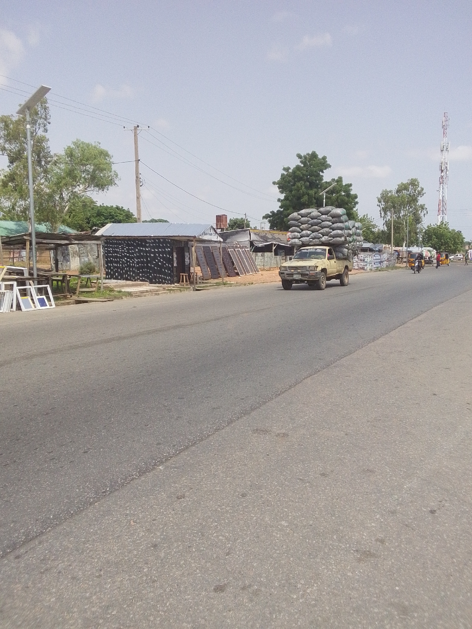 The image shows a street scene in Jalingo, Nigeria. A light-colored pickup truck is driving on the right side of the road, heavily loaded with sacks of what appears to be charcoal or grain. The truck is positioned in the foreground, slightly to the right of center. On the left side of the road, there are several market stalls or shops with various goods displayed, including what look like window frames or shutters. There are also some trees and utility poles visible. In the background, a tall telecommunications tower can be seen. The sky is overcast with some clouds. The overall impression is of a busy, somewhat dusty roadside environment.